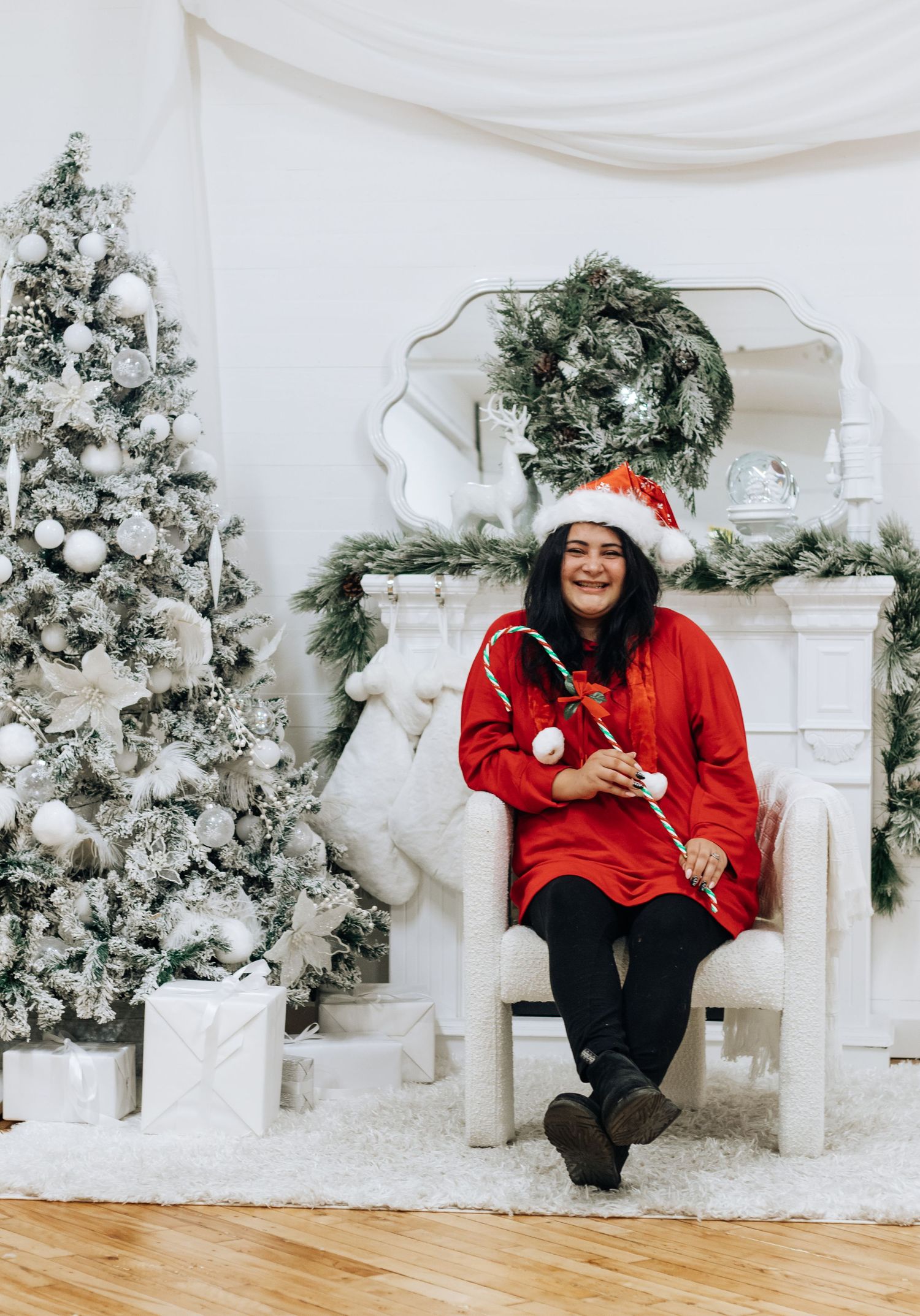A festive holiday photo shoot setup with white Christmas trees, decorative fireplace and person in red Santa outfit.