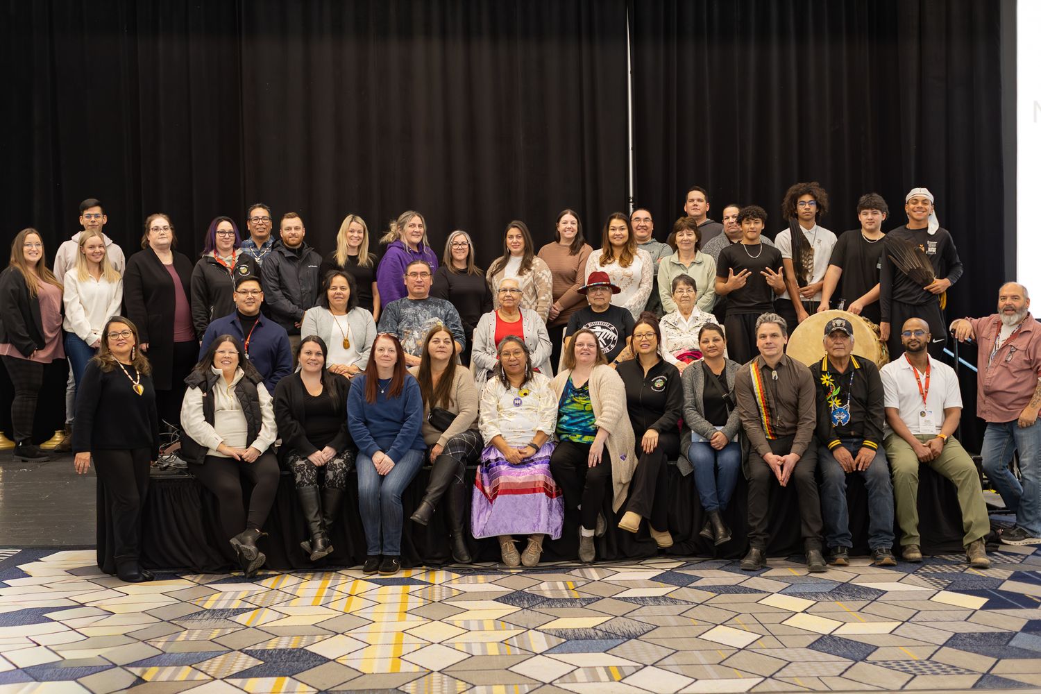Large group photo of event attendees seated together against a black curtain backdrop.