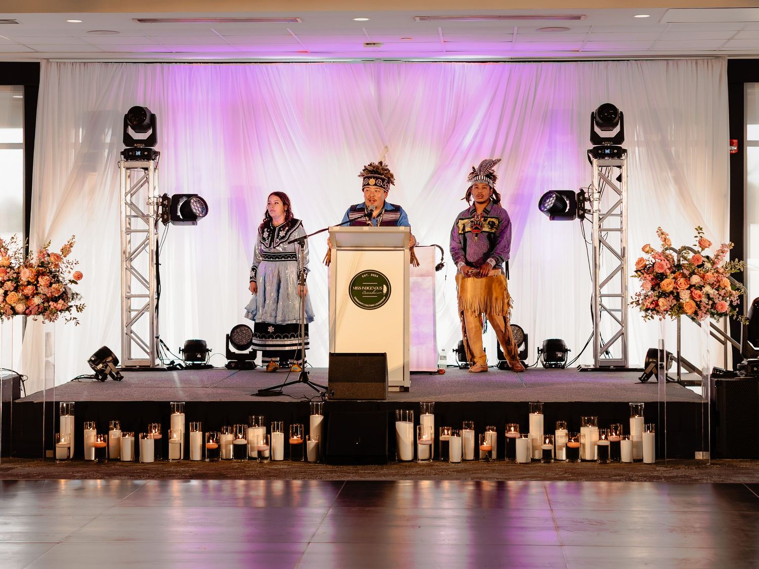 Three people on stage at formal event, center speaker at podium in traditional Indigenous headdress.