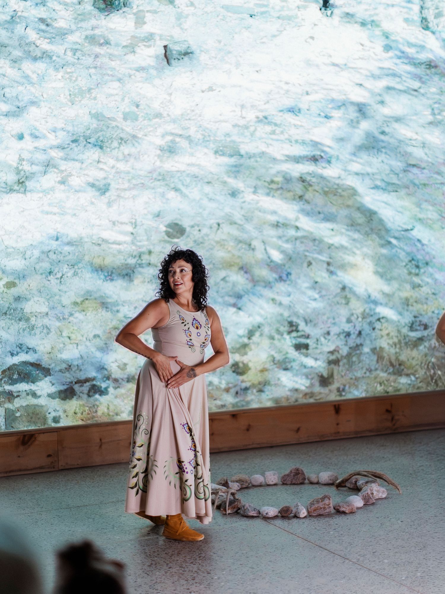 Woman in floral maxi dress stands before large aquatic mural with stone circle arrangement on floor.