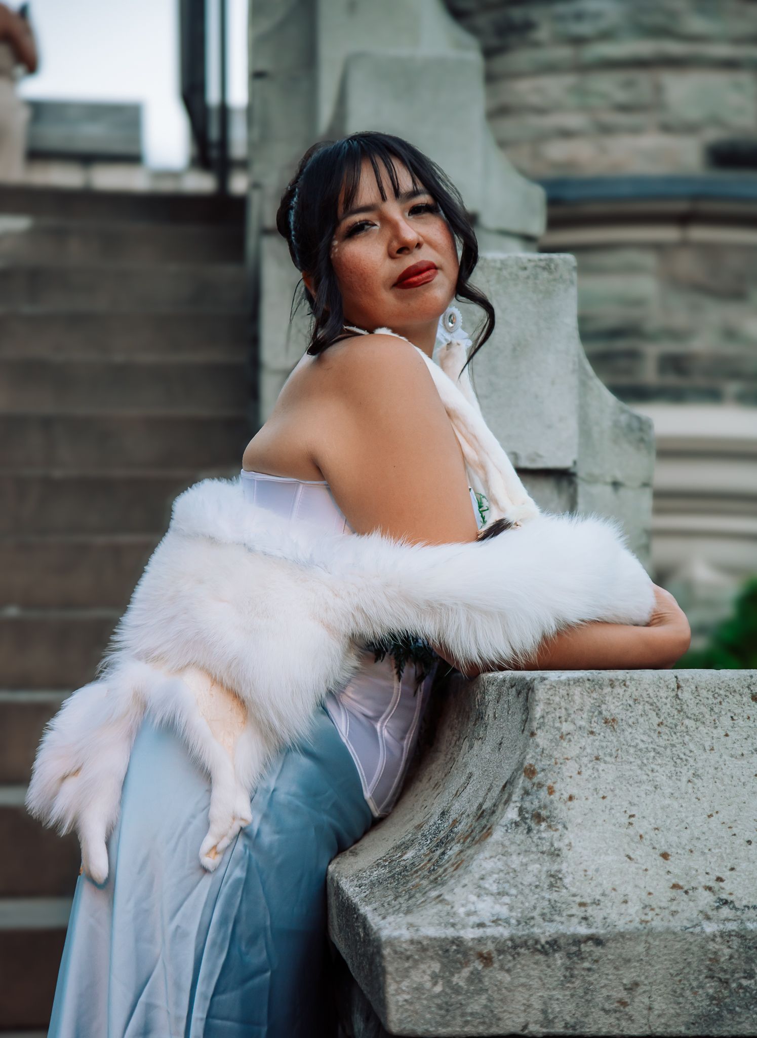 Elegant woman in white fur stole and blue gown poses on stone steps of a historic building.