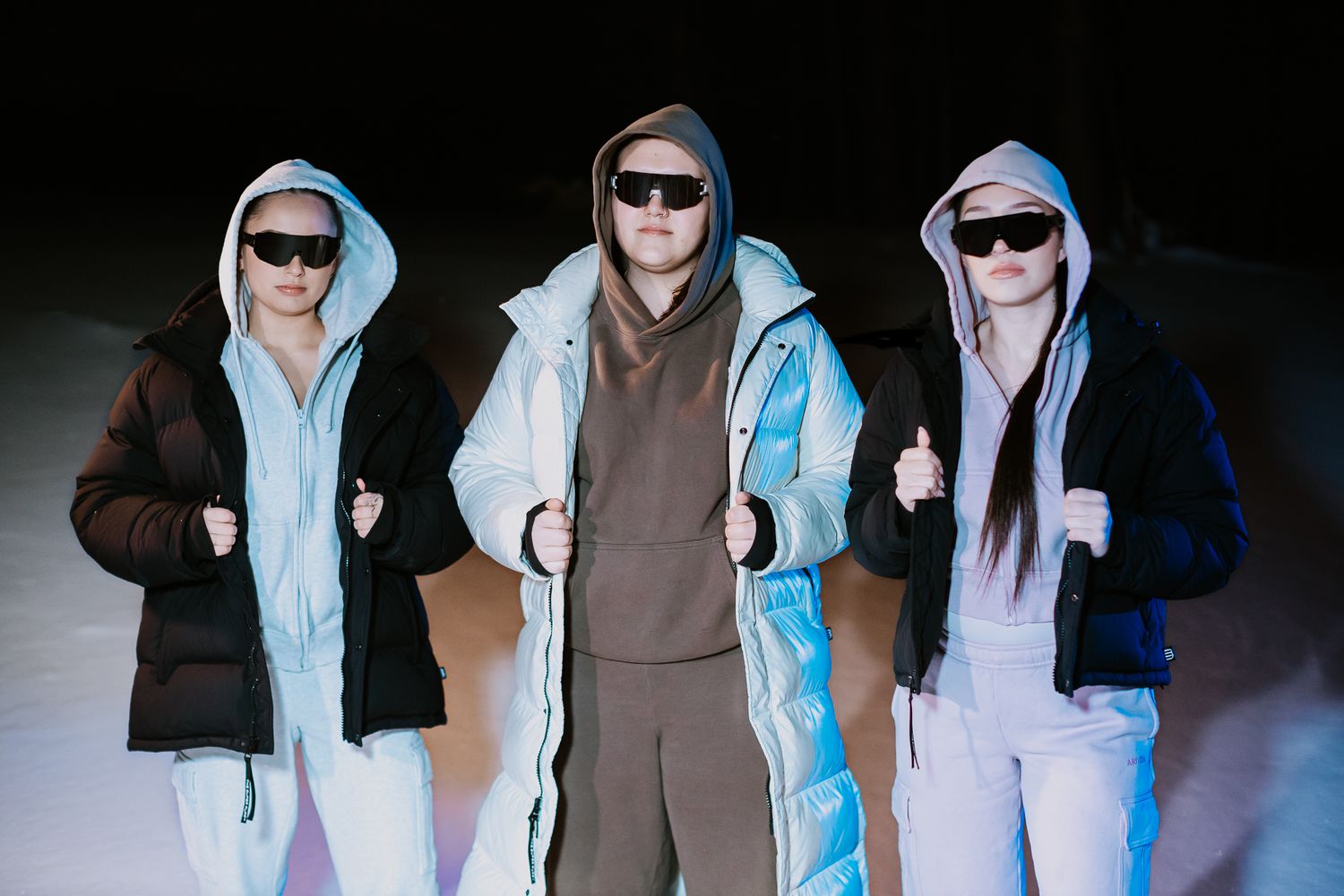 Three models wearing hooded puffer jackets and sunglasses posed together against a dark background.