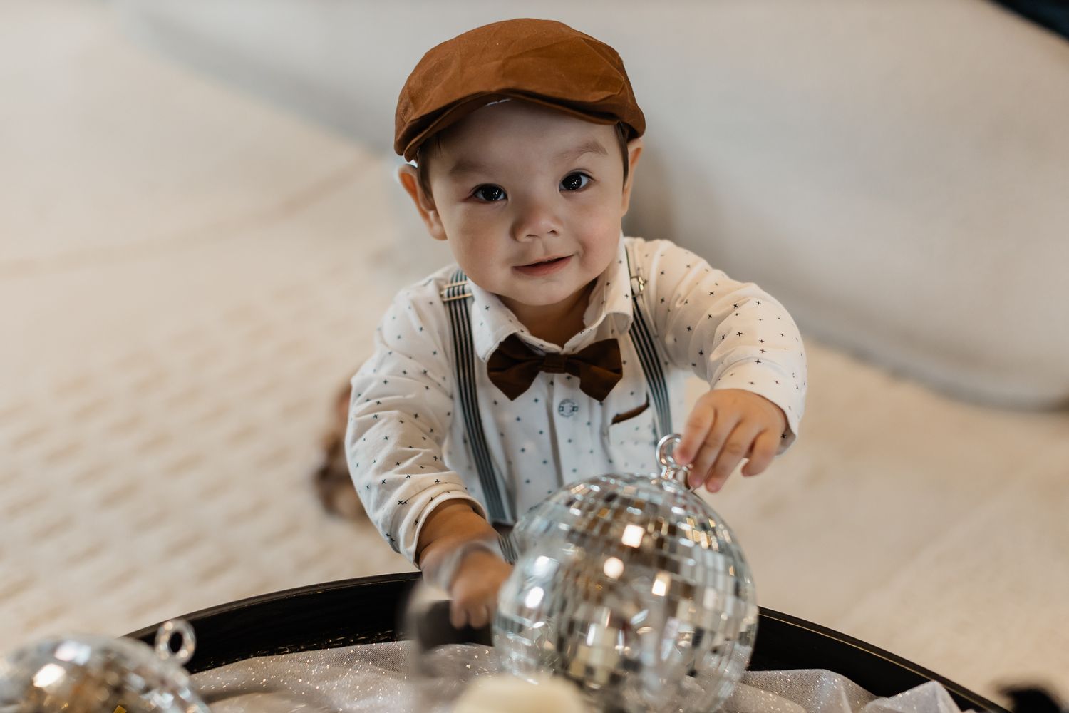 Adorable baby boy in brown cap and bow tie smiling while playing with silver disco balls.