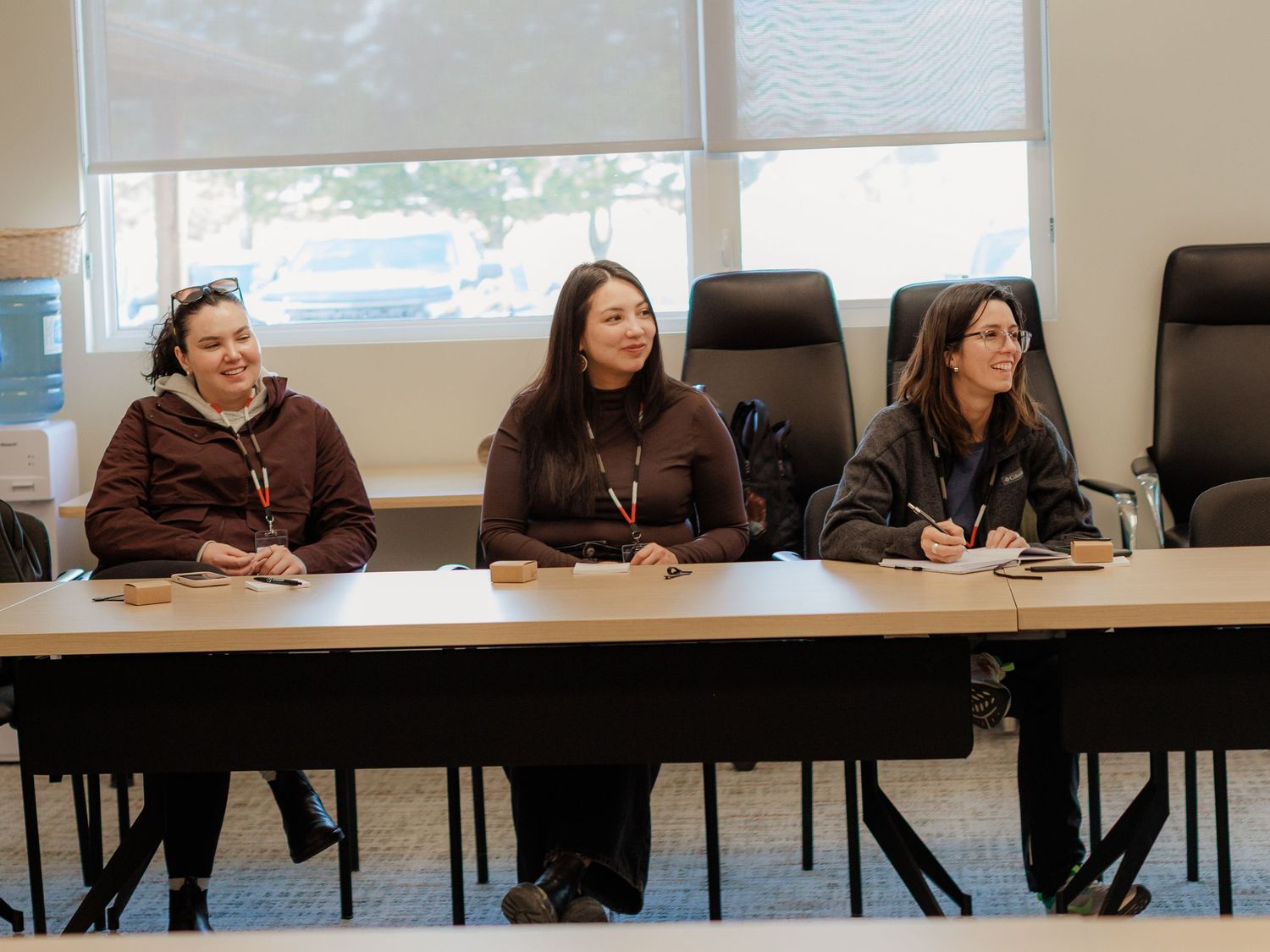 Three women sitting at a conference table with notepads, smiling during a professional meeting in a modern office.
