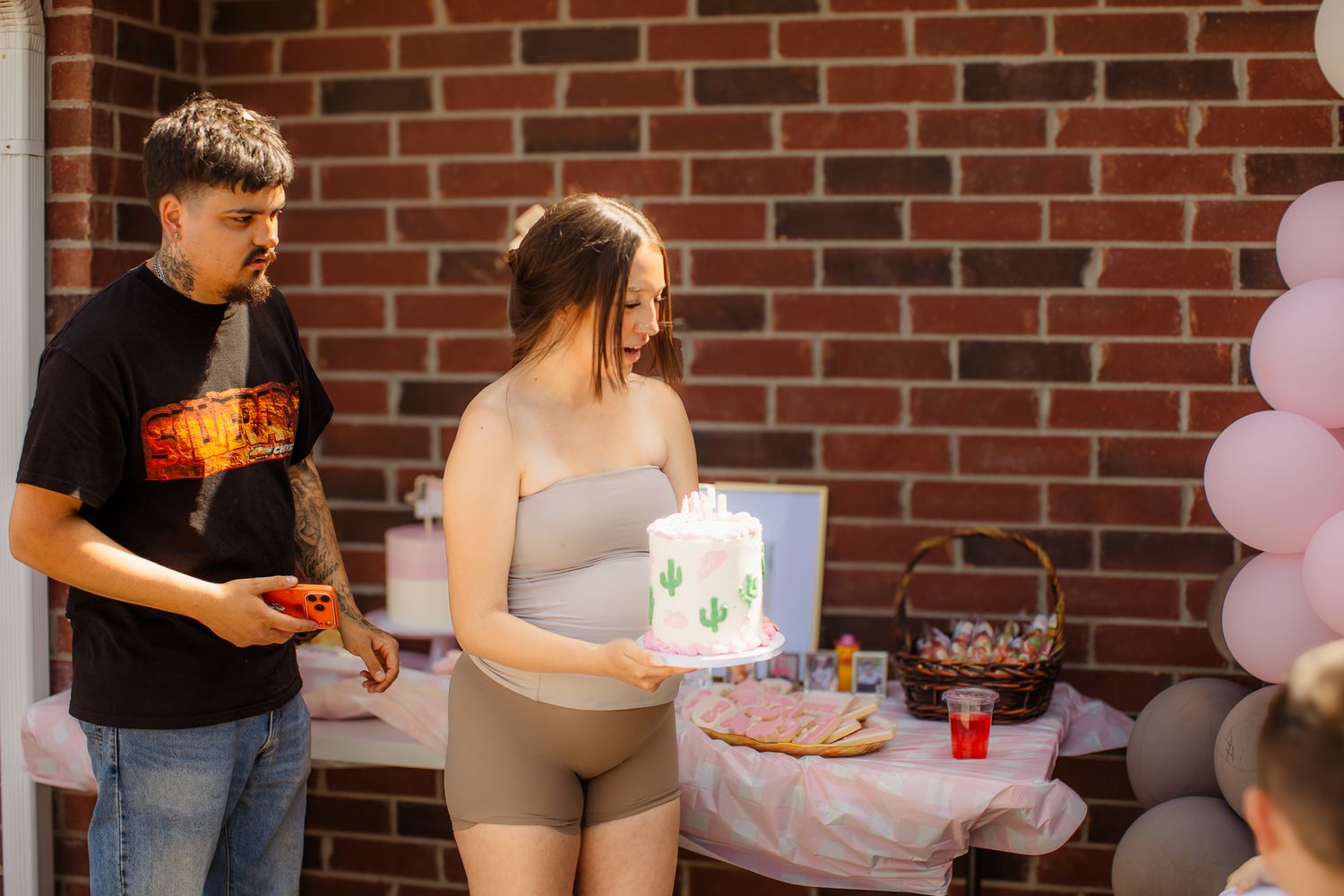 Pregnant woman holding white cactus cake at baby shower party with man beside her near balloon decorations.