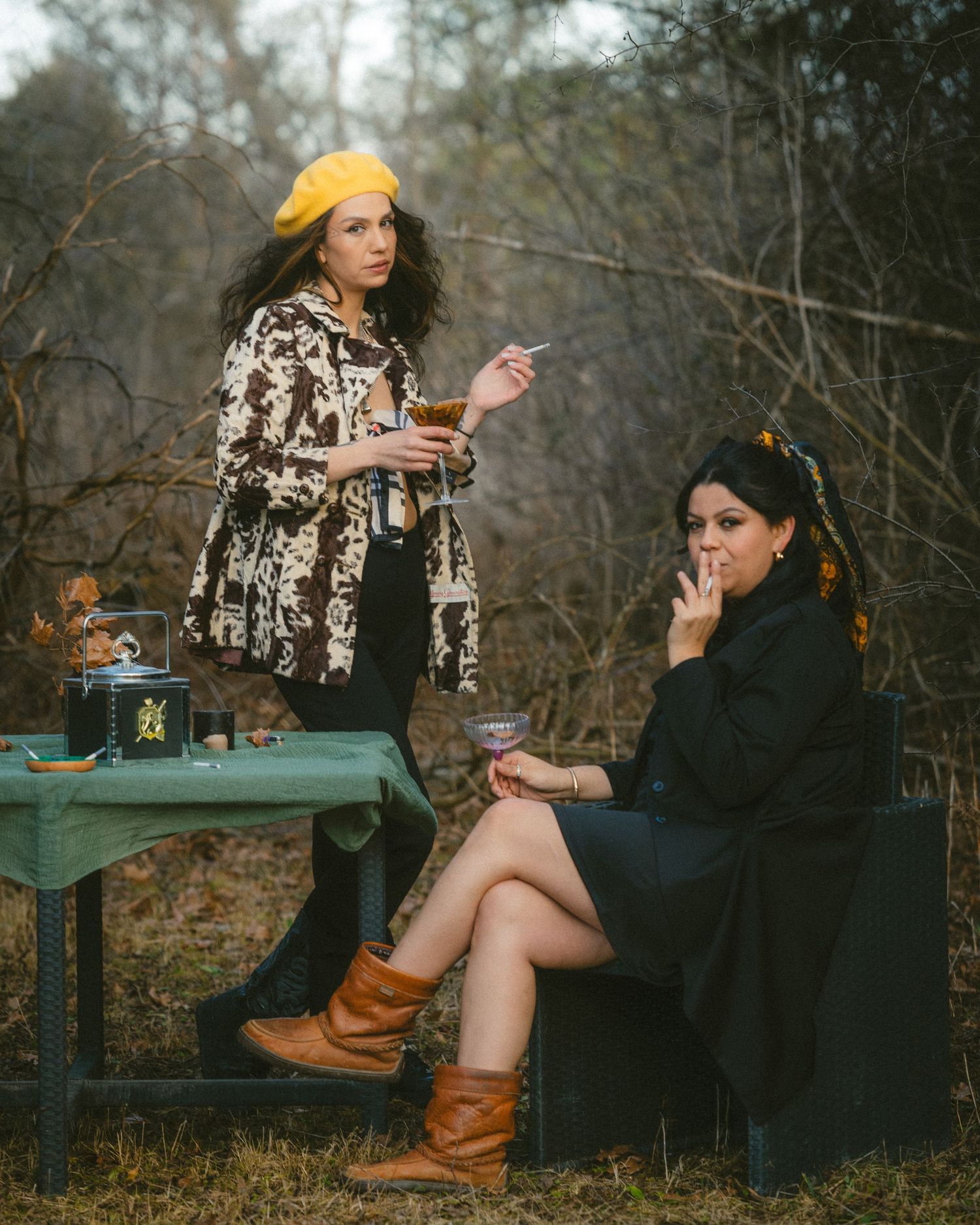 Two stylish women pose outdoors with cigarettes, one in yellow beret and cow-print jacket, other in black coat holding wine glass.