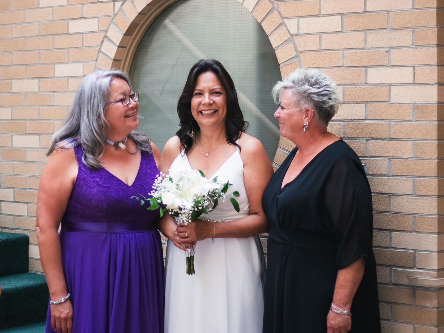 Bride in white gown holding bouquet stands with two women in purple and black dresses outside a brick church.
