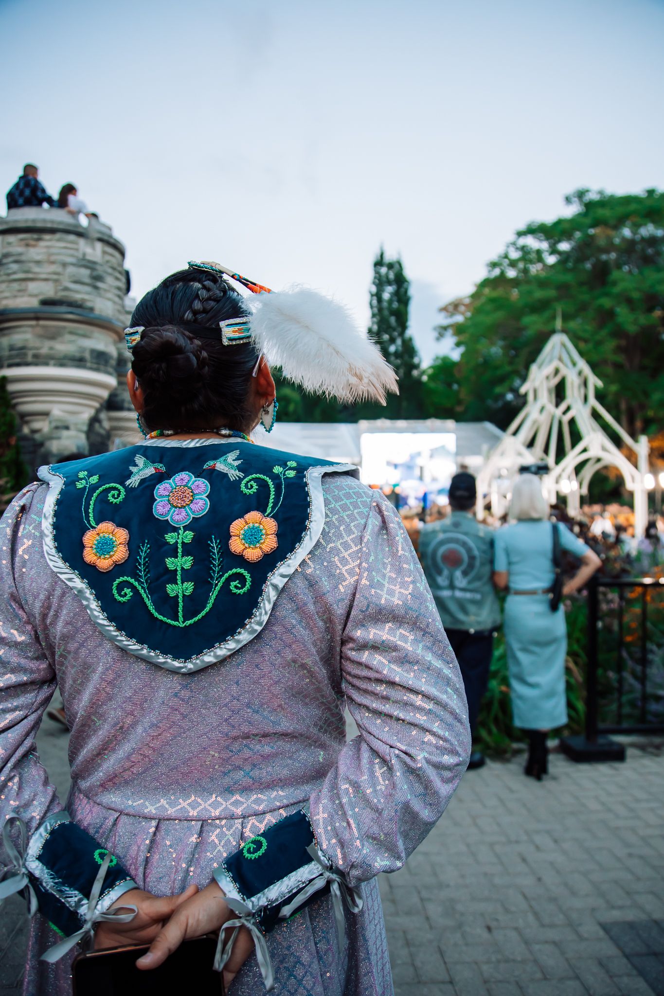 Person in traditional Indigenous regalia with floral beadwork and feather headdress at an outdoor cultural event.