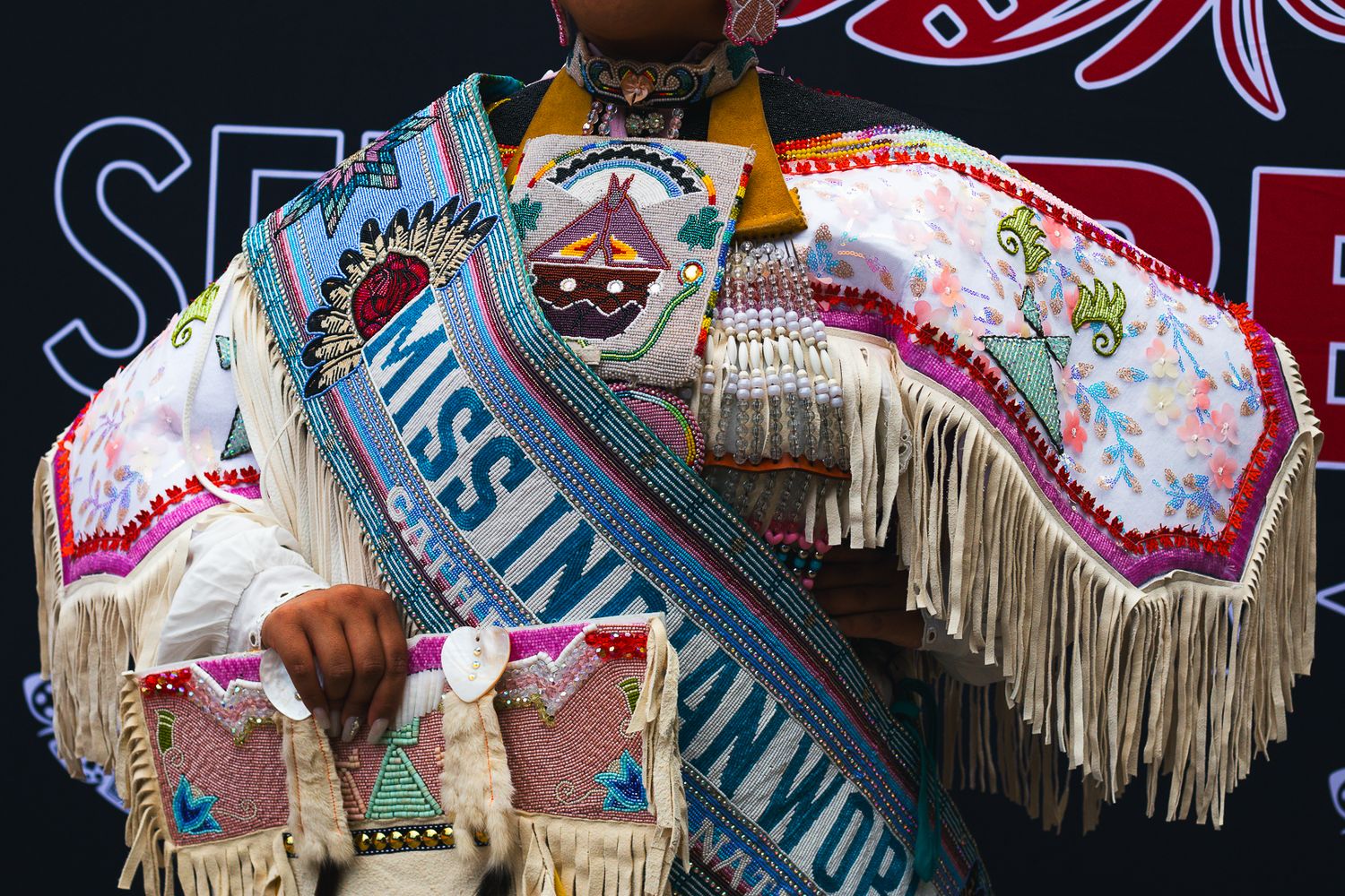 Woman wearing Miss Indian World sash and traditional beaded regalia with fringe at a cultural event.
