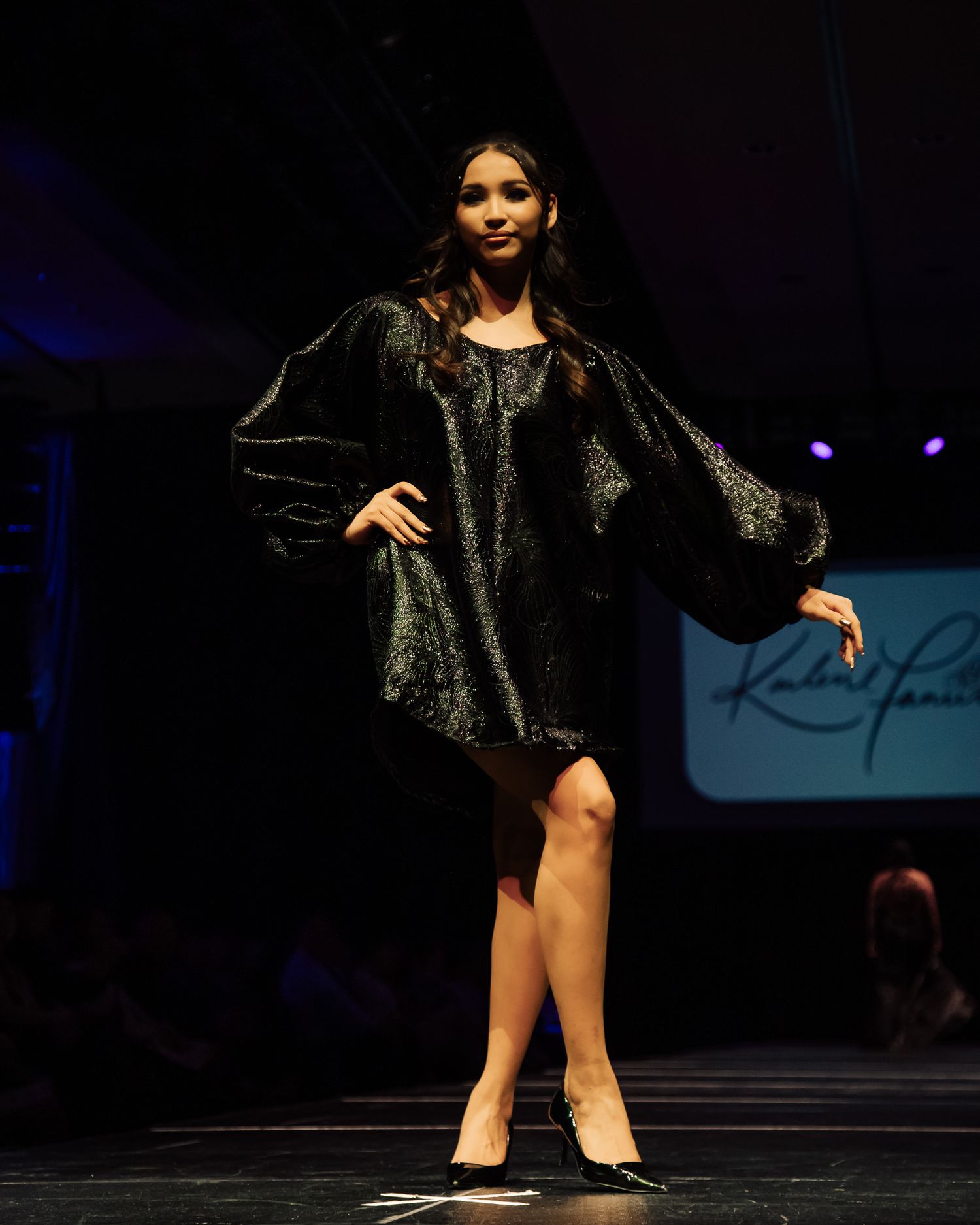 Model walks runway in black sequin mini dress with dramatic wide sleeves and black heels at fashion show.