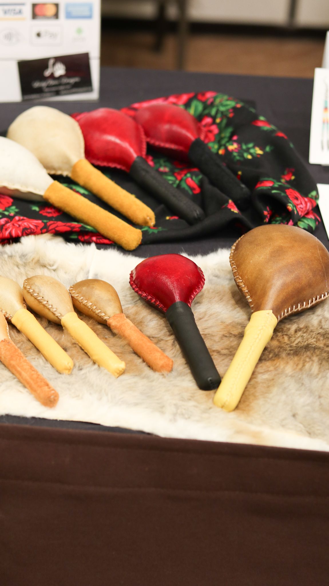 Handcrafted leather maracas and wooden percussion instruments displayed on fur and floral fabric at a cultural market.