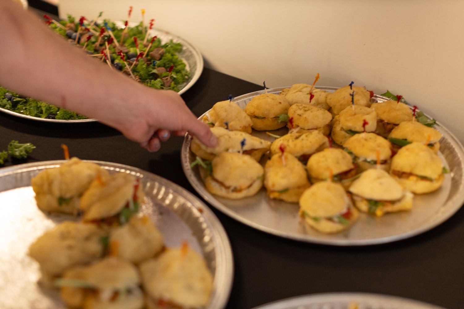 Hands reaching for small sandwich appetizers arranged on silver serving platters.