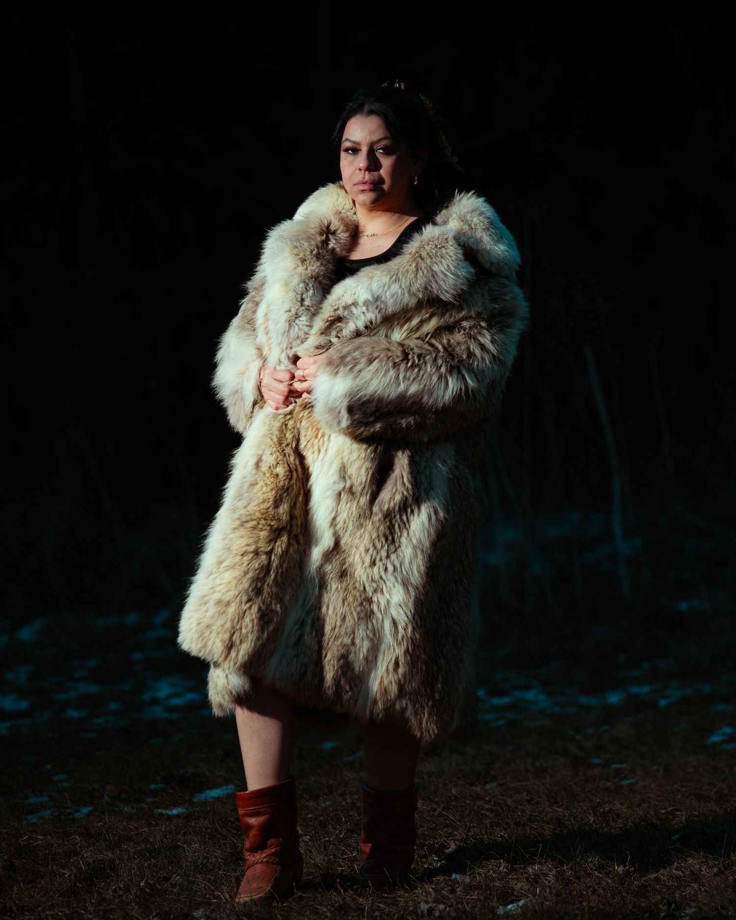 Woman in a large fur coat and brown boots stands dramatically in a dark outdoor setting.