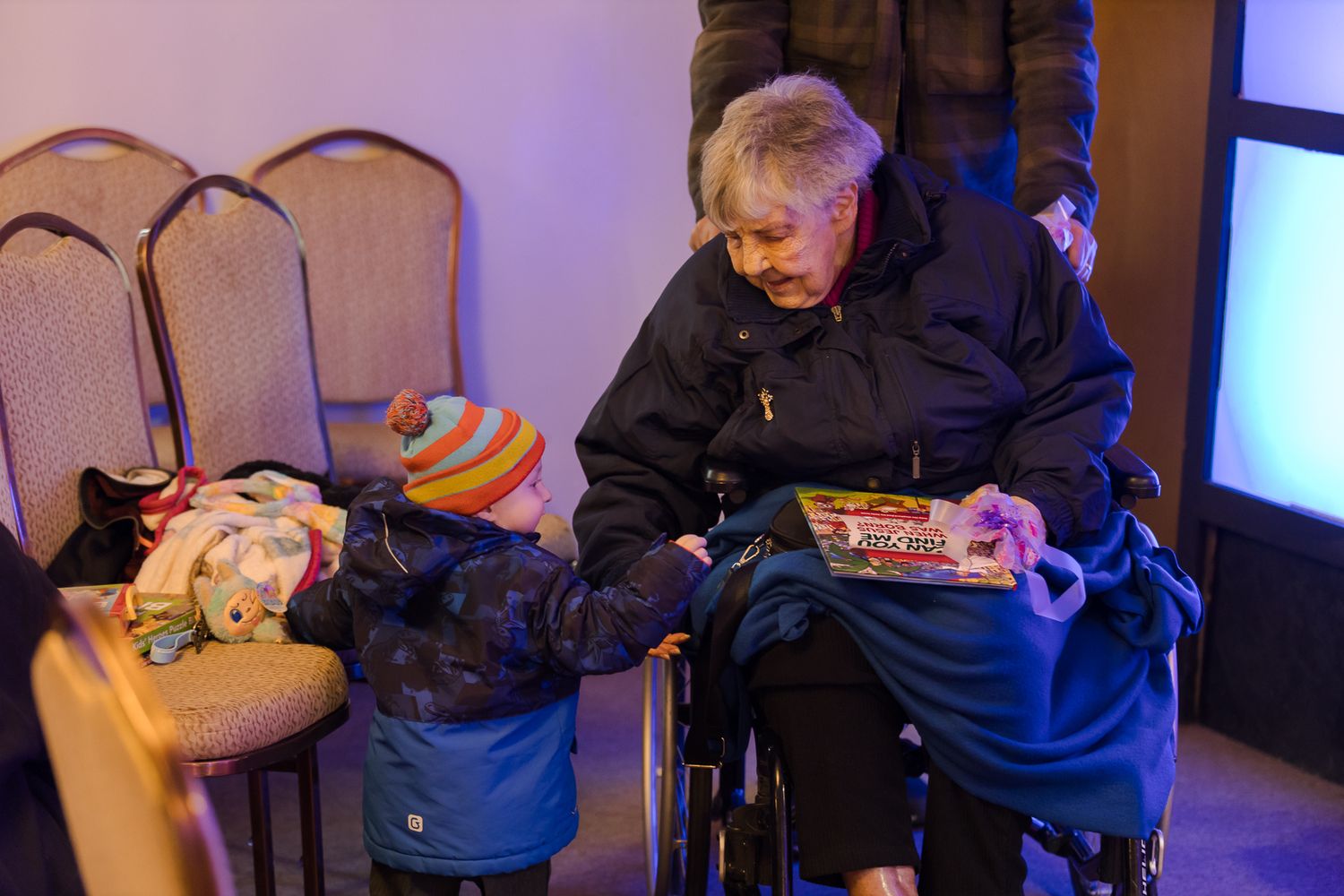 Elderly person in wheelchair sharing a moment with a young child in a colorful hat indoors.