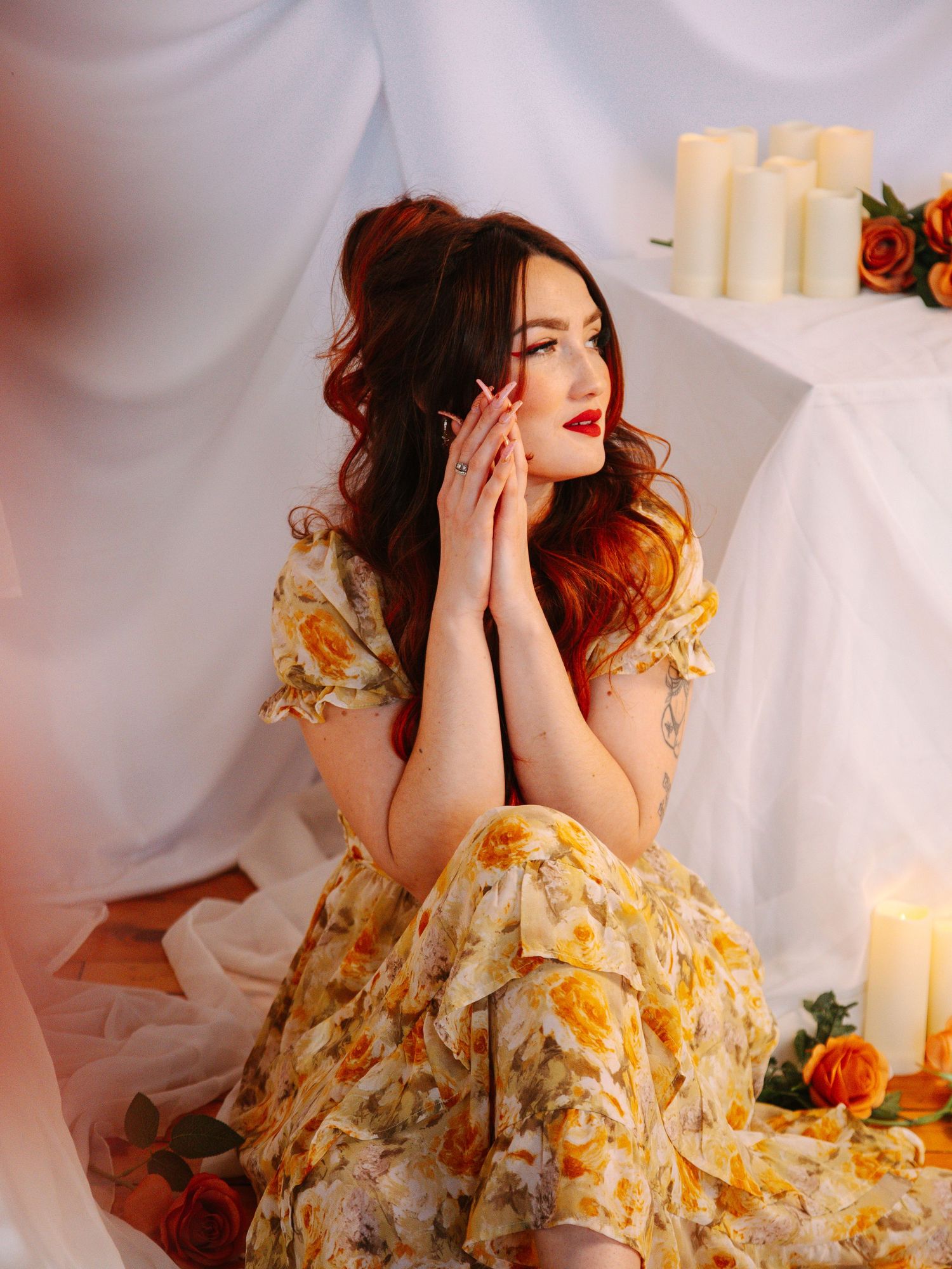 Woman in yellow floral dress with red hair poses thoughtfully among candles and orange roses against white draping.