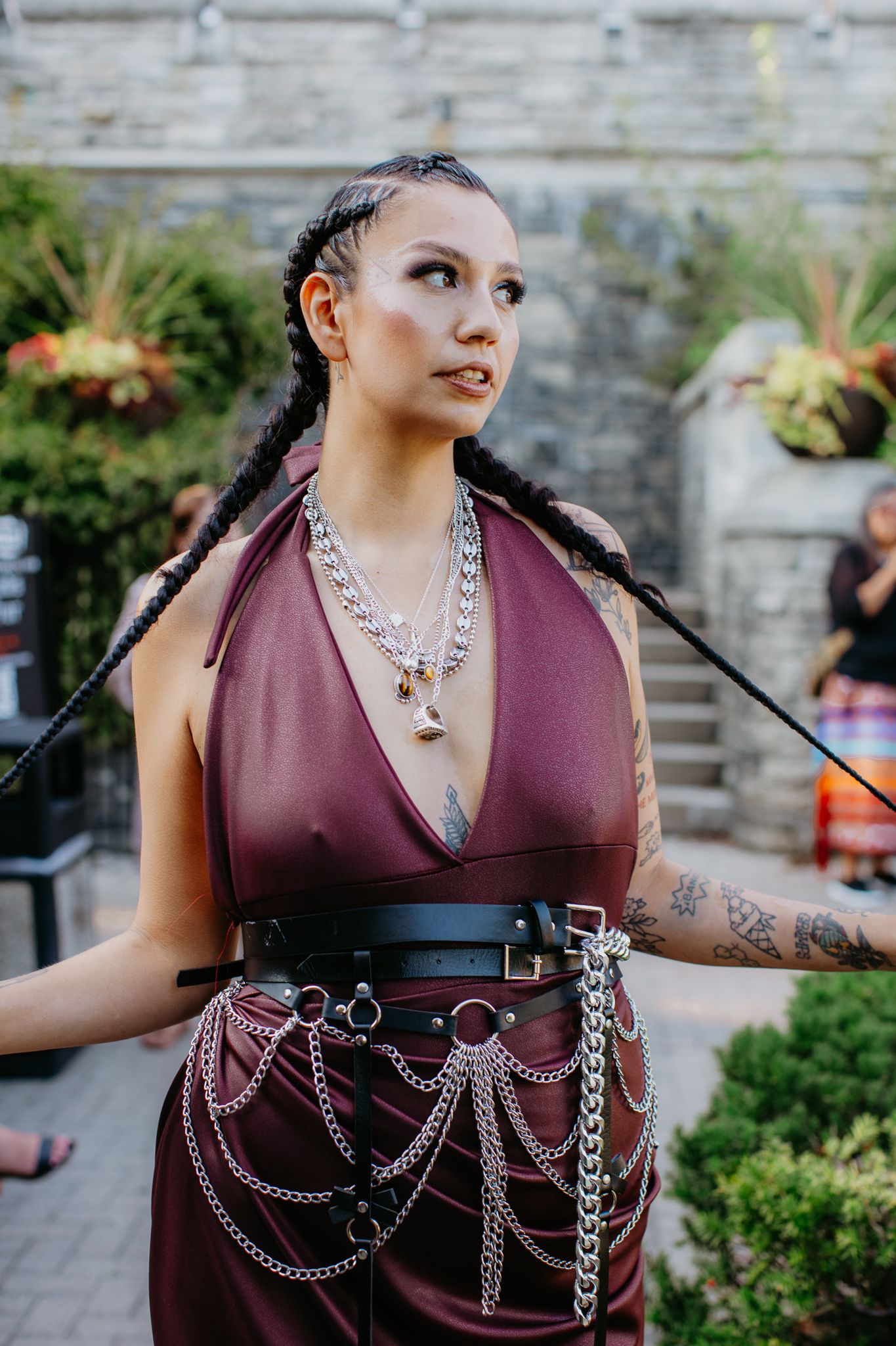 Woman in burgundy halter dress with chain belt, silver necklaces, long braids, and tattoos posing outdoors.