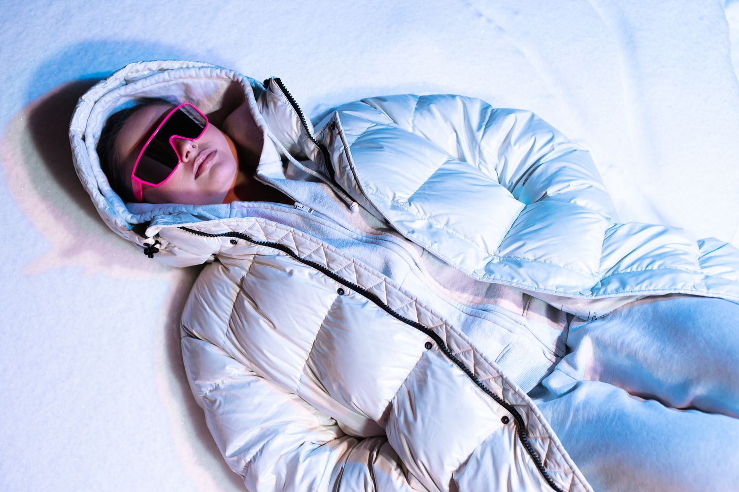 Woman lying in snow wearing a shiny white puffer jacket, hood, and pink shield sunglasses.