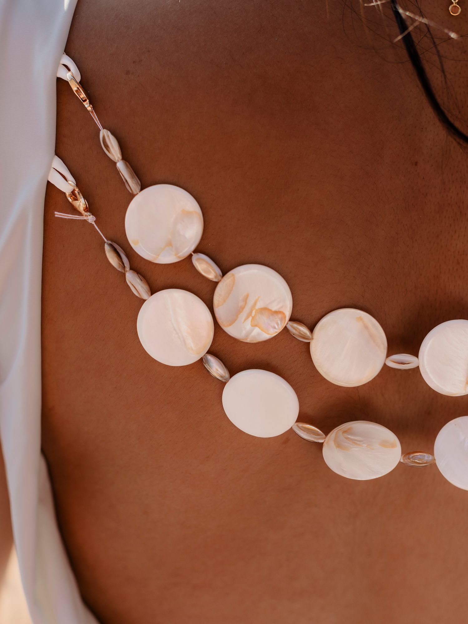 Close-up of layered shell necklaces with round white mother-of-pearl discs and small cowrie shells on tanned skin.
