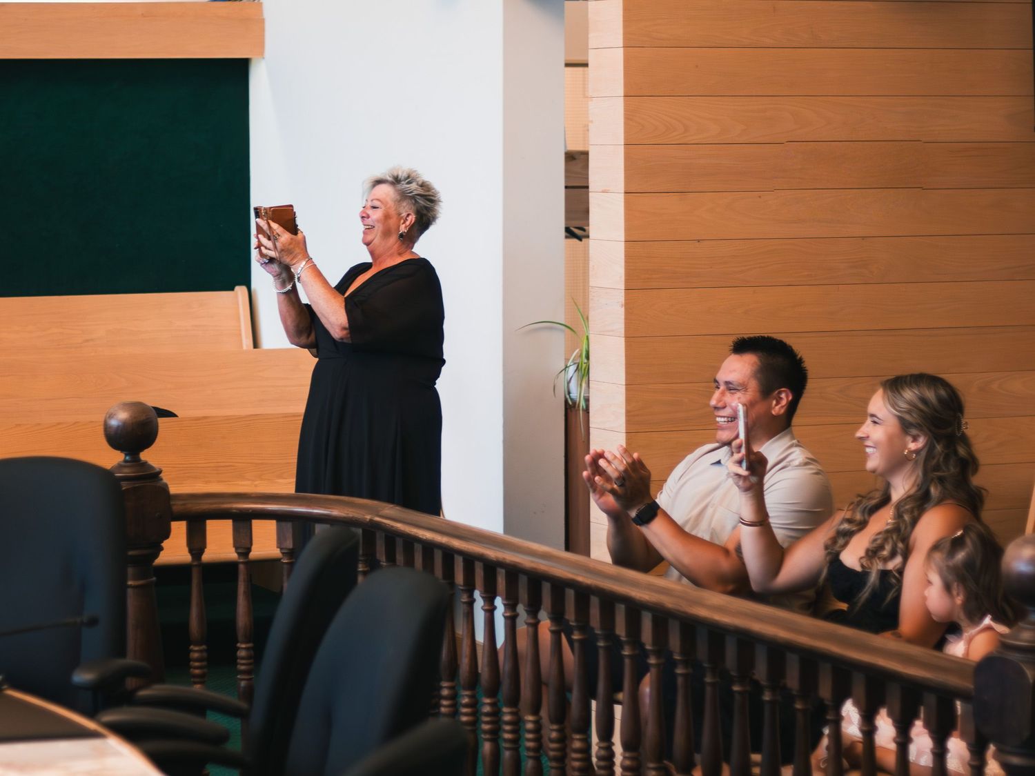 Woman in black dress reads at podium while guests applaud at indoor wedding ceremony balcony.
