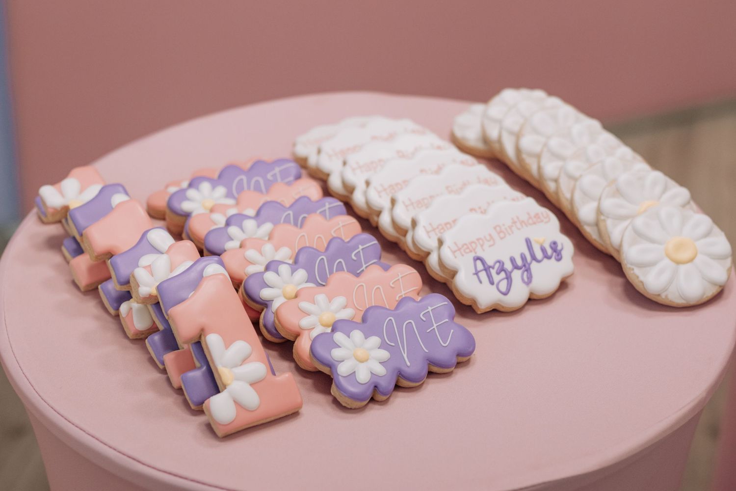 Decorated sugar cookies in purple and pink with floral designs displayed on a pink table.