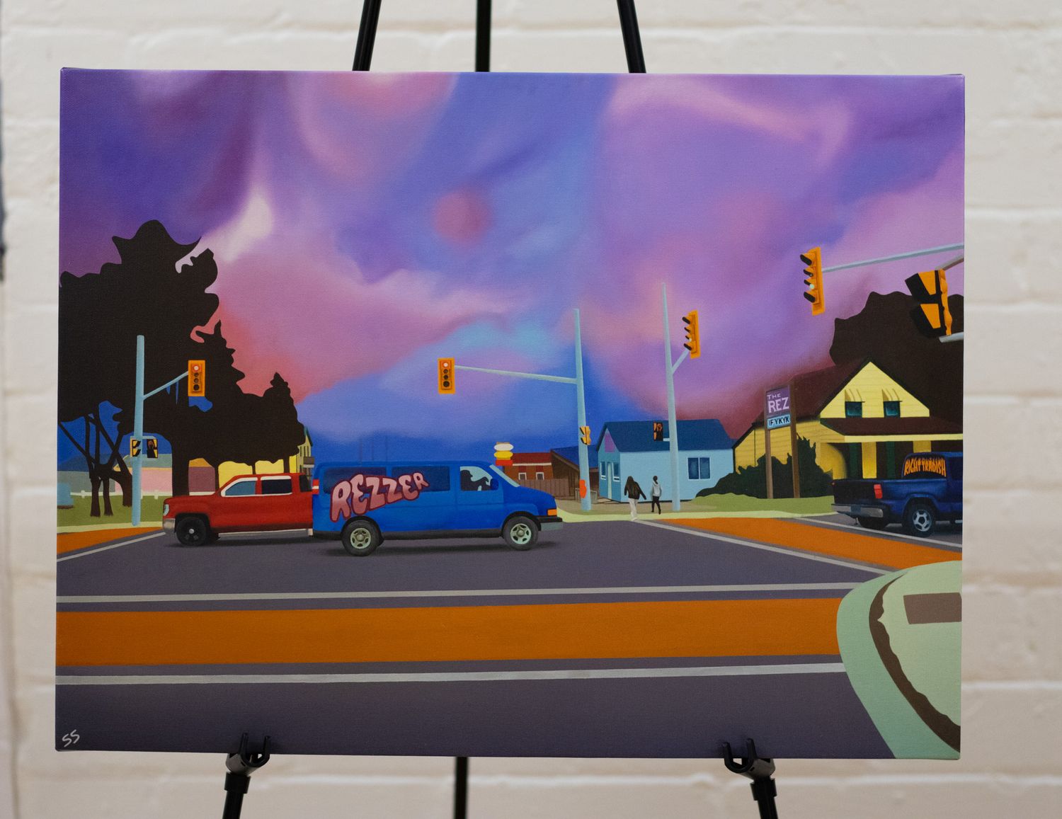 Colorful sunset cityscape artwork displayed on easels against white brick wall.