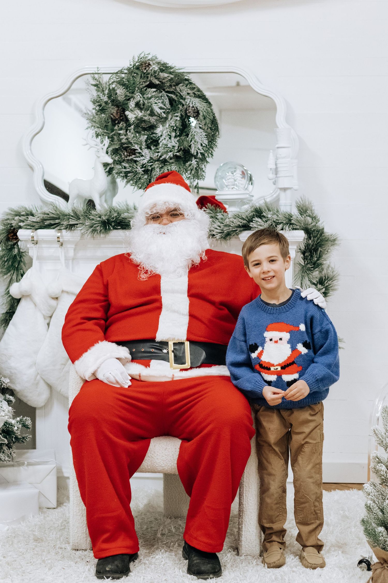 Child sits with Santa Claus in front of a white Christmas backdrop with evergreen decorations.