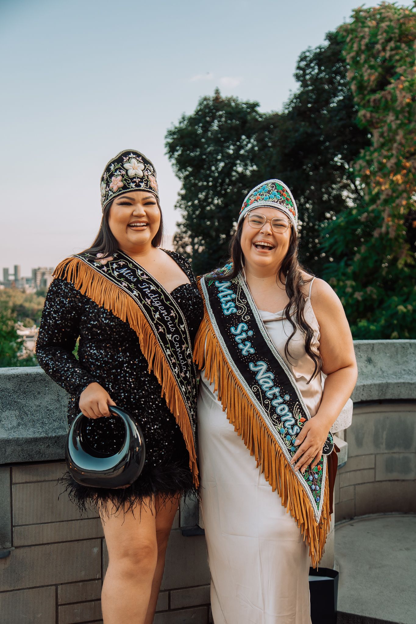 Two women wearing Indigenous regalia, sashes, and beaded crowns smile outdoors at a Miss Six Nations pageant event.