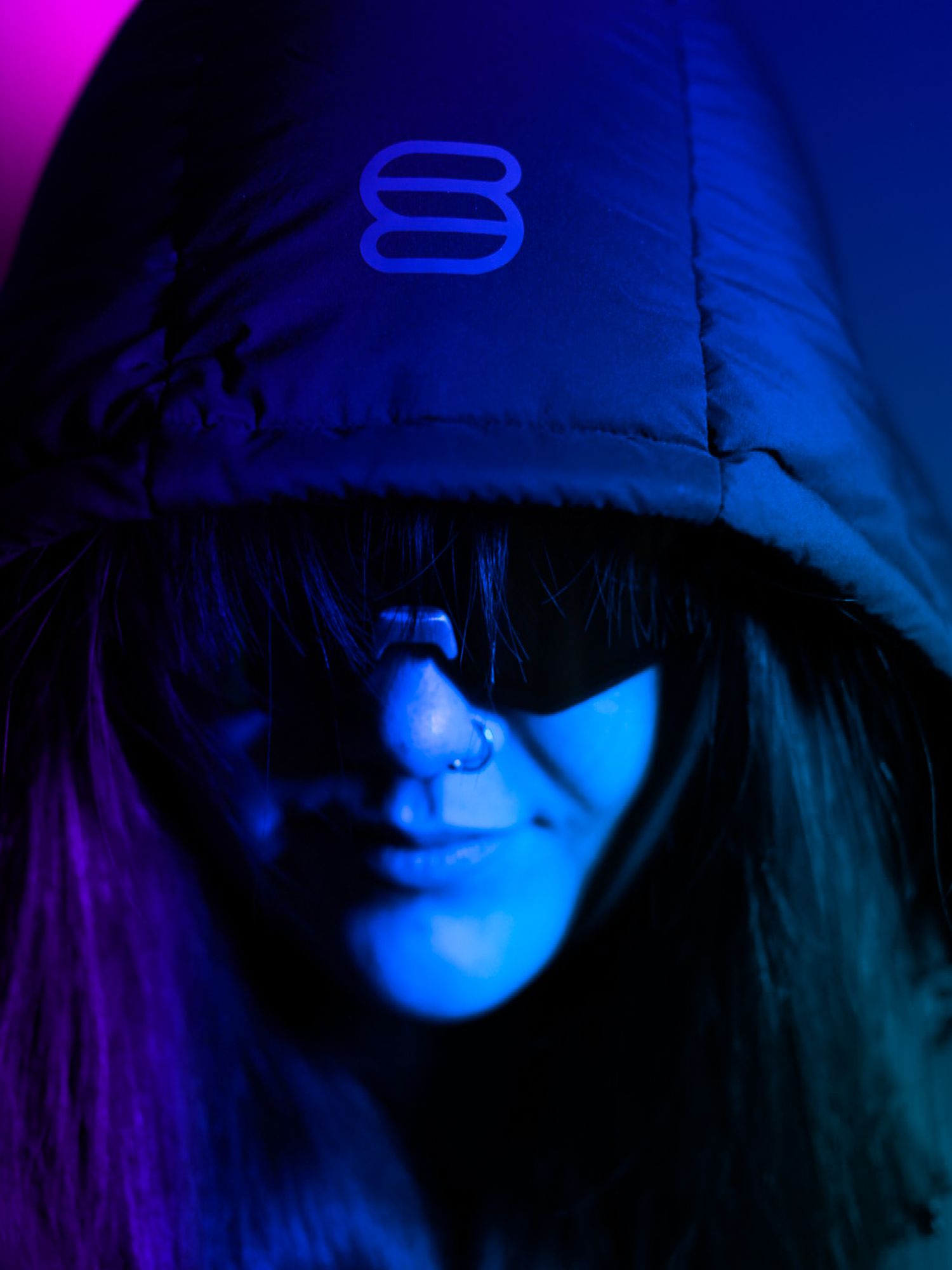 Person wearing a blue hooded puffer jacket with logo and dark sunglasses under dramatic blue and purple neon lighting.