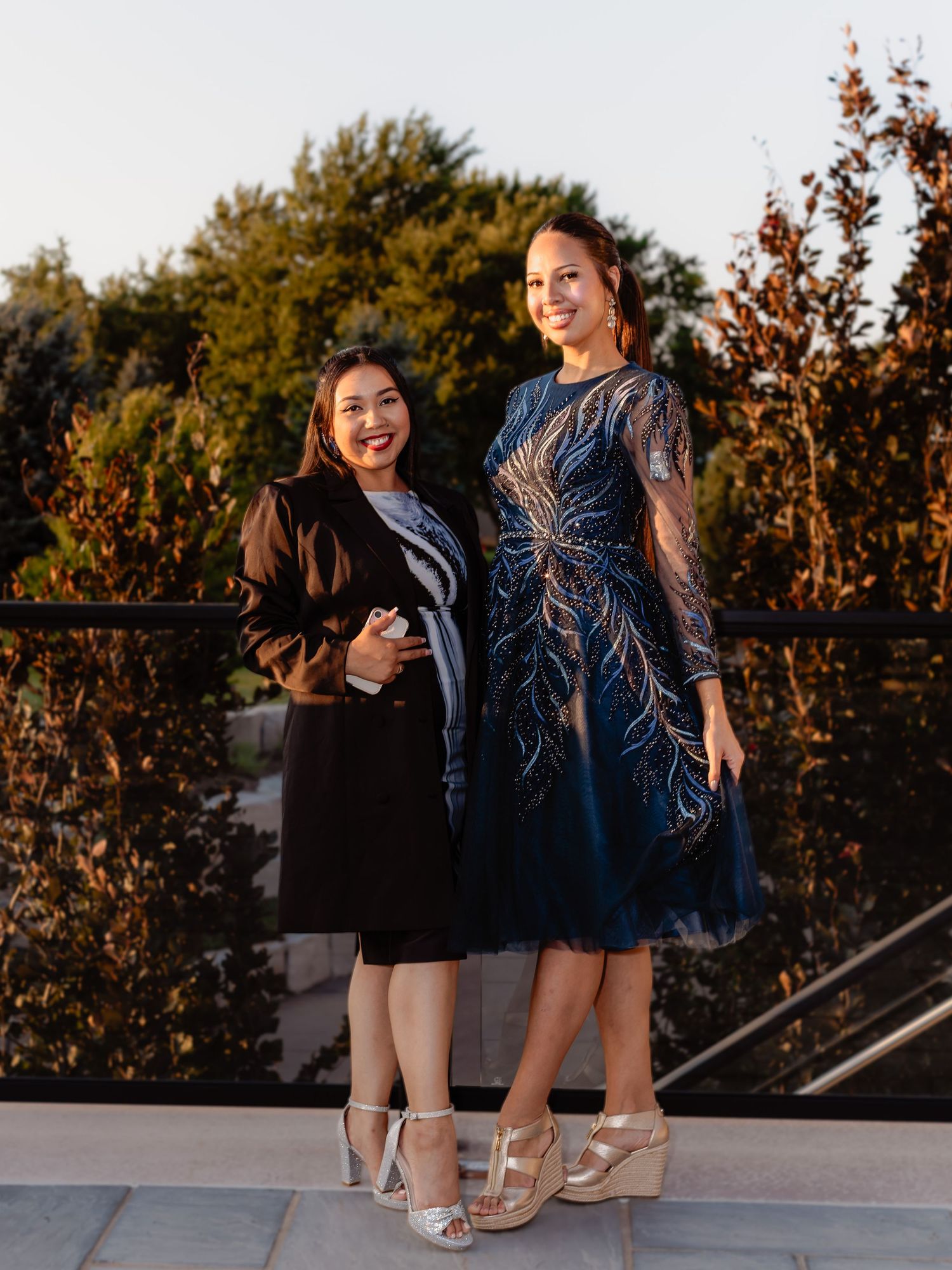 Two women in elegant formal dresses posing together on an outdoor terrace at golden hour.