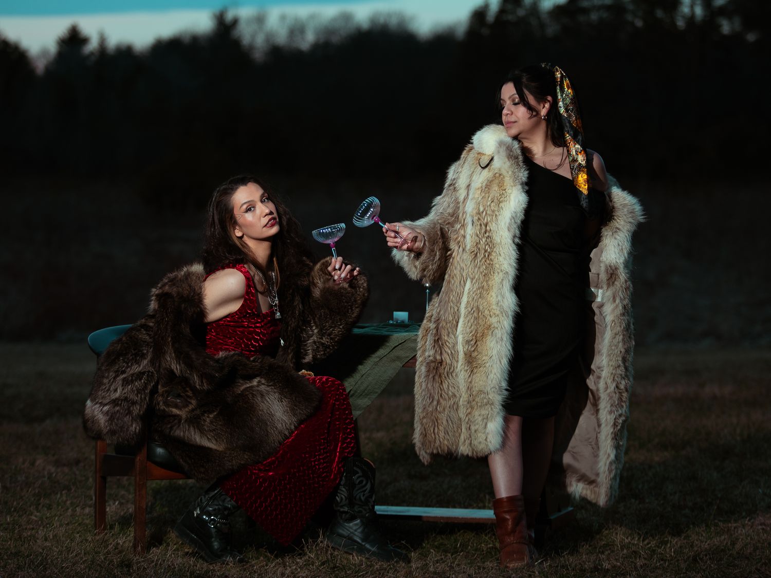 Two women in fur coats holding cocktail glasses in dramatic outdoor evening setting.