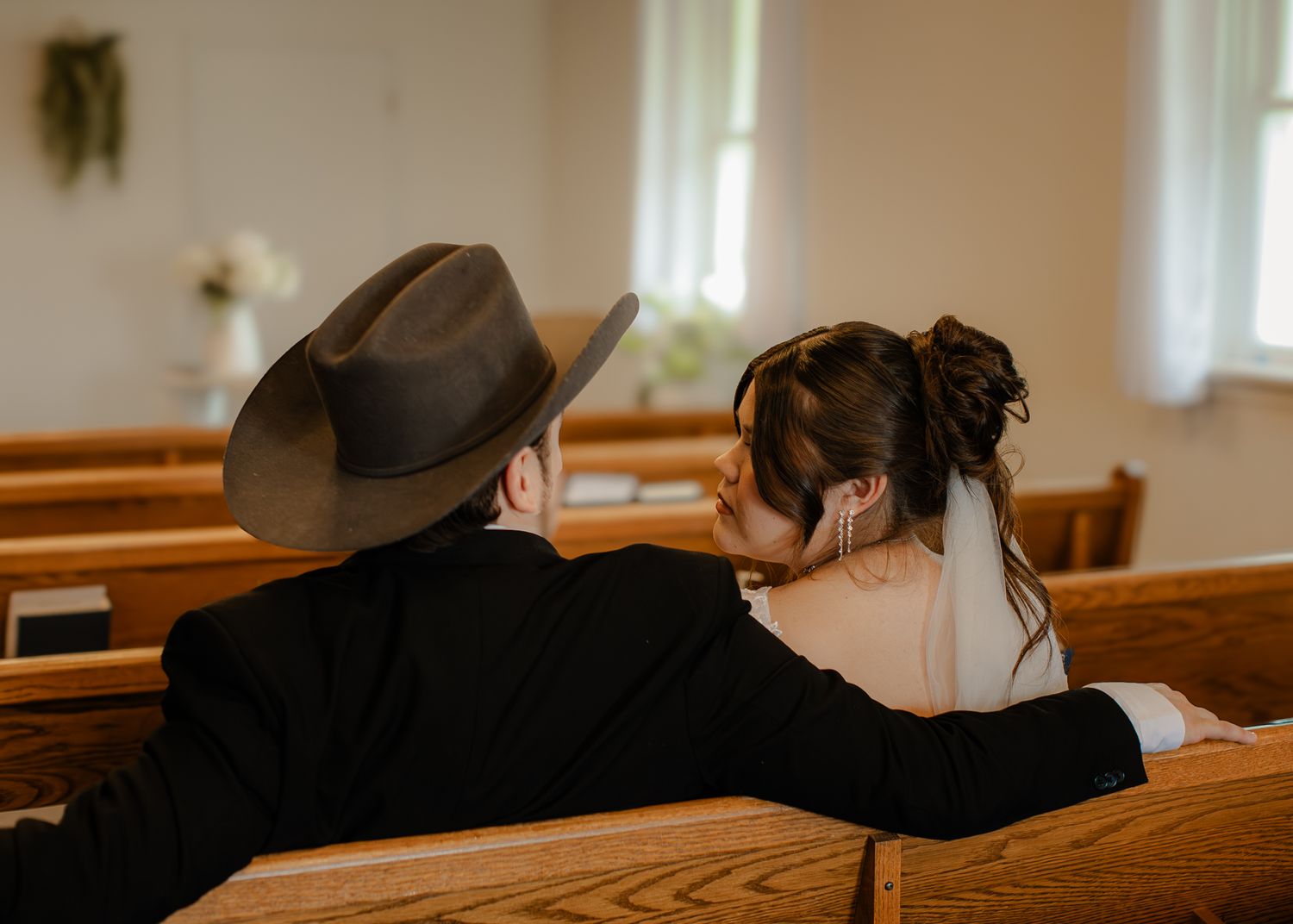 Bride and groom in cowboy hat sit intimately together on church pew during wedding ceremony.