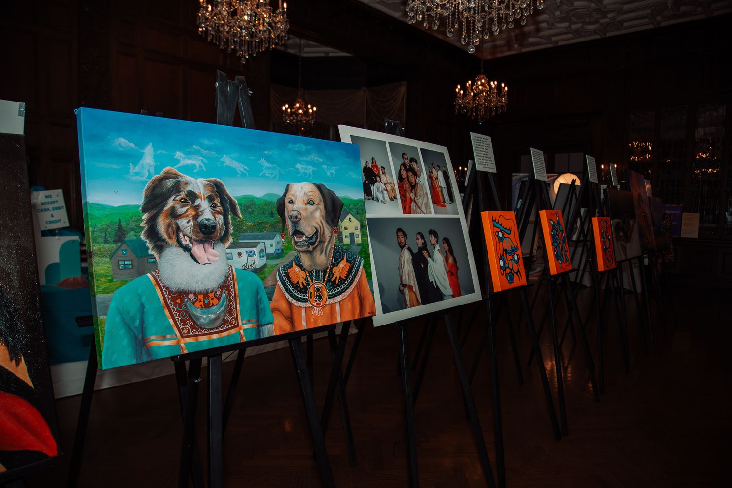 Gallery display of colorful dog portrait paintings on easels in a dimly lit chandelier ballroom.