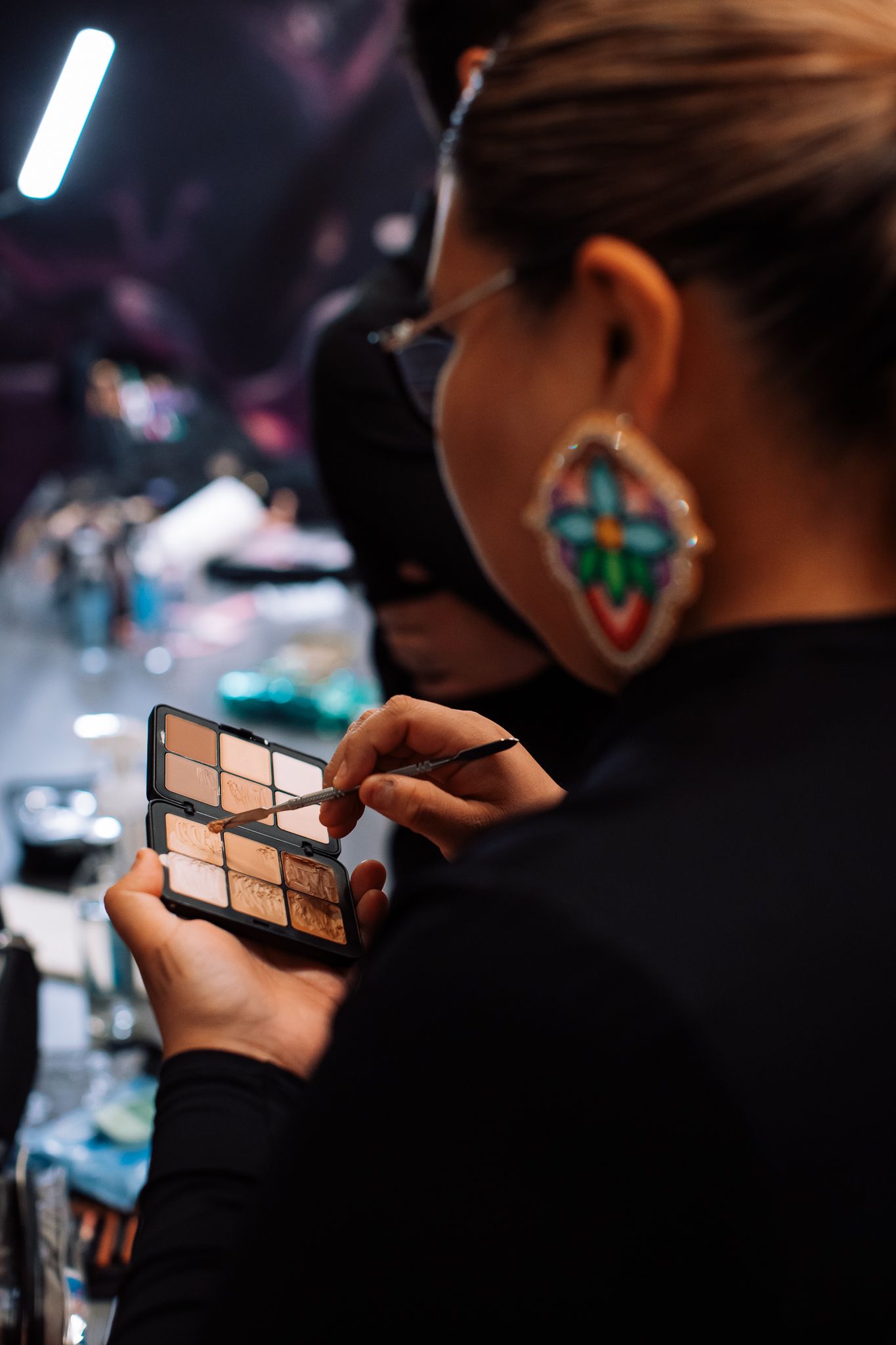 Makeup artist holding an eyeshadow palette with a brush, wearing decorative earrings in a professional studio setting.