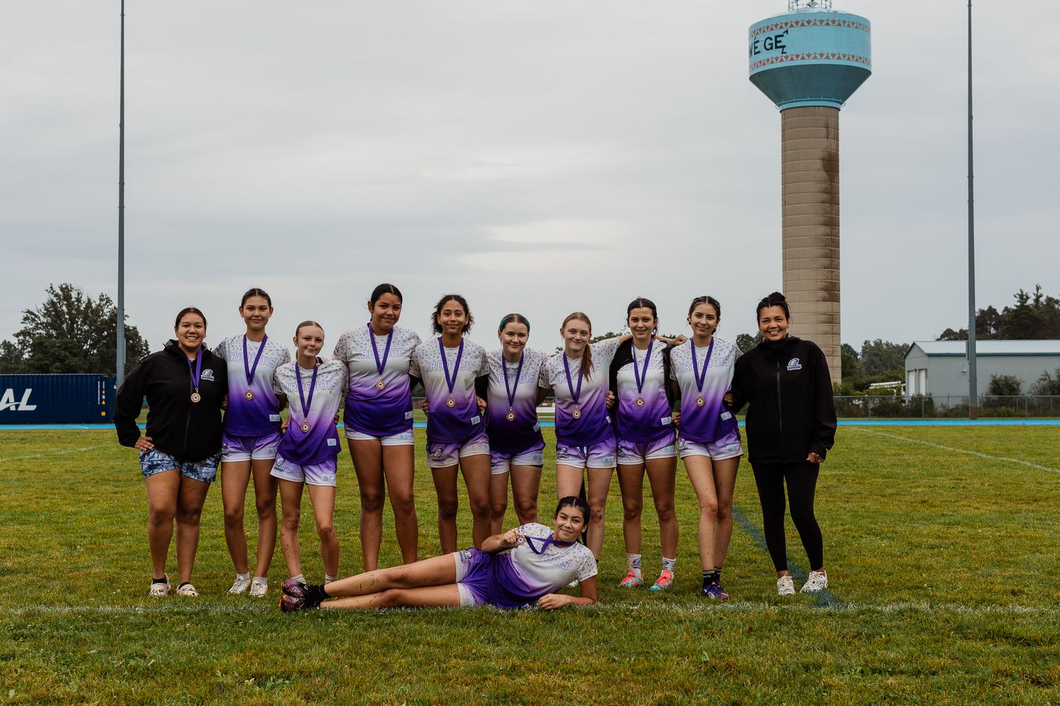 Girls cheerleading team in purple uniforms posing with medals on a sports field with a water tower in the background.