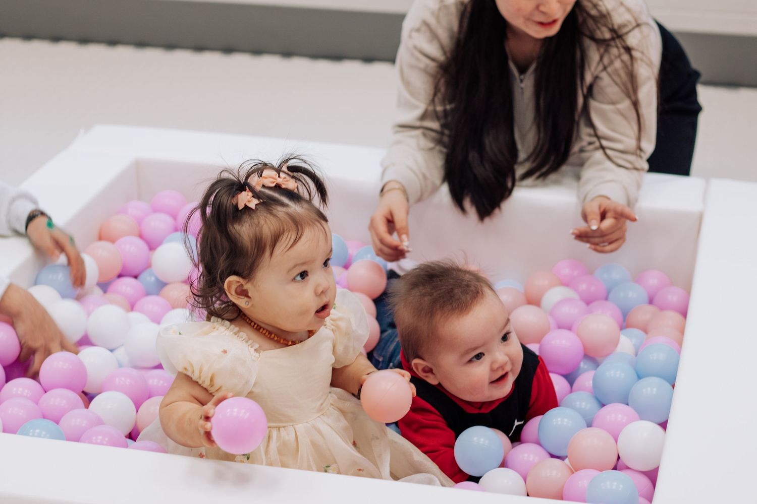 Two babies playing in a colorful ball pit with pastel balls, supervised by a woman leaning over the edge.