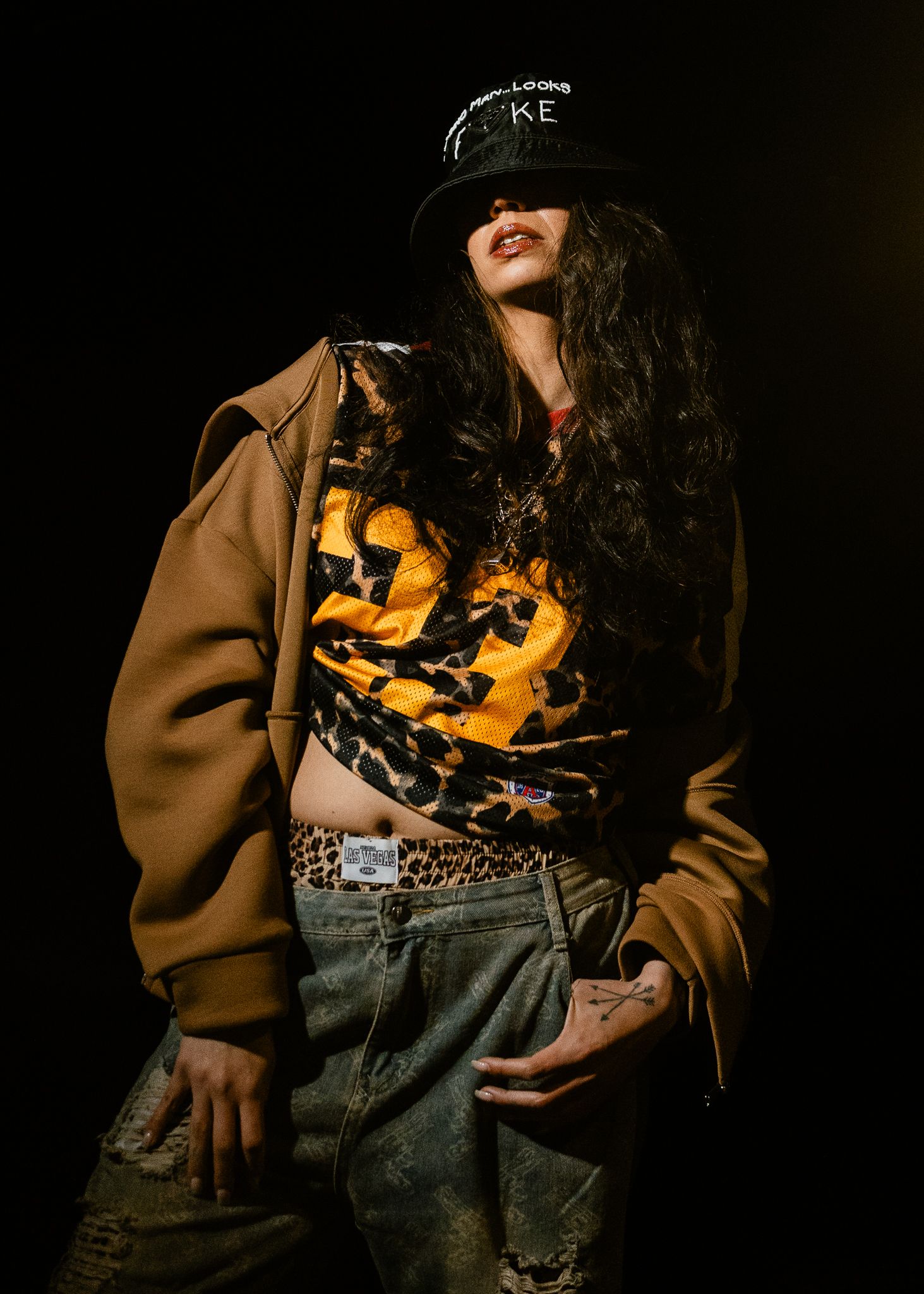 Woman in leopard print crop top, brown oversized jacket, distressed jeans, and black cap poses confidently.