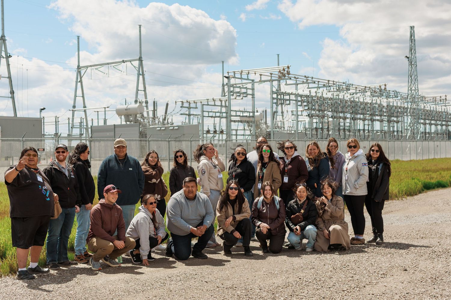 Large group of professionals posing outdoors in front of an electrical substation on a cloudy day.