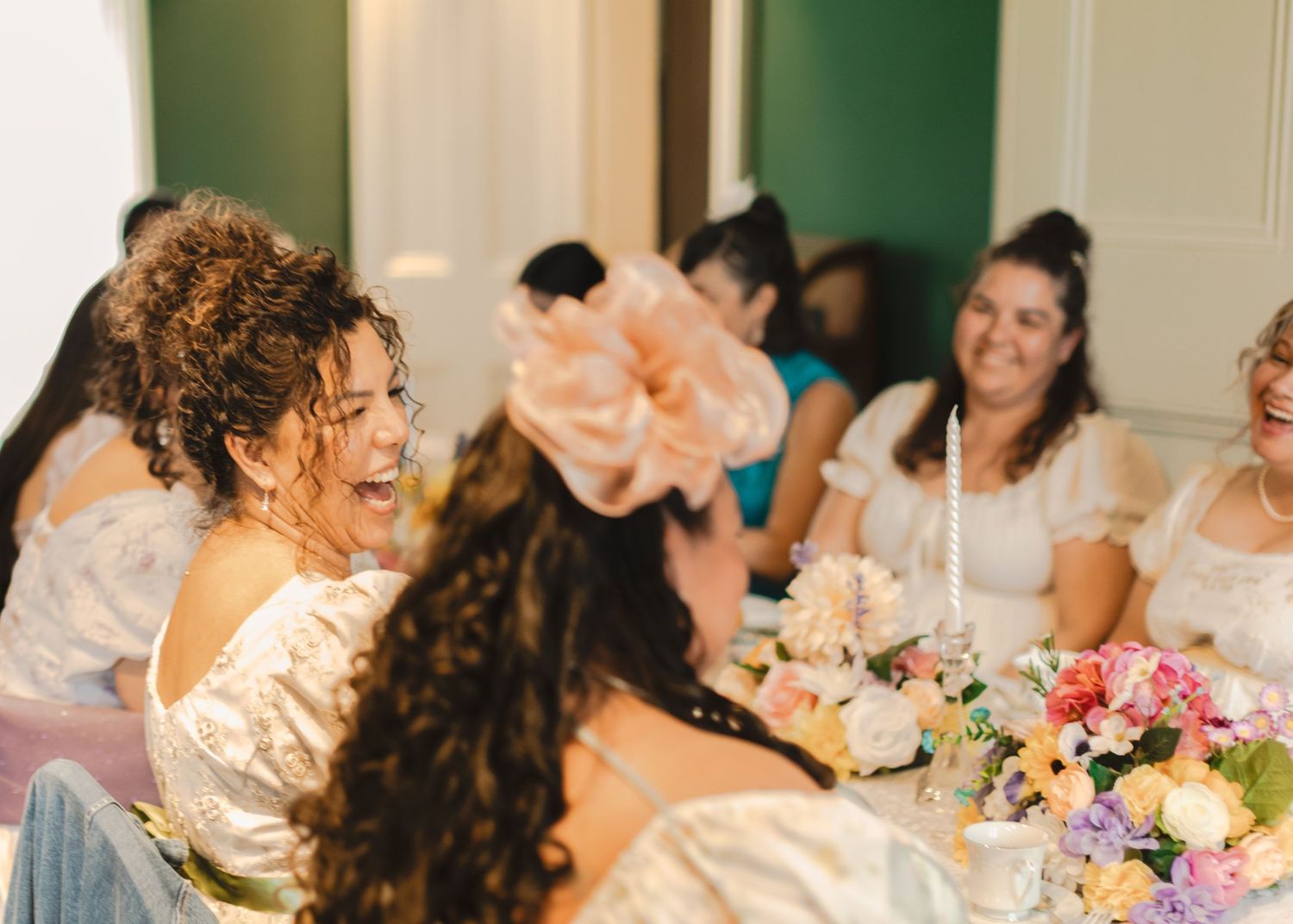 Women in white dresses laughing at a floral-decorated table during a bridal party celebration.