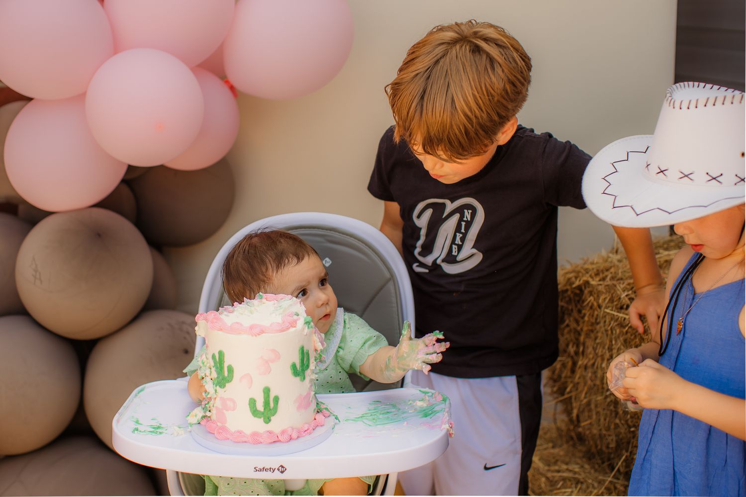 Baby smashing first birthday cake at cactus-themed party while two children watch nearby.