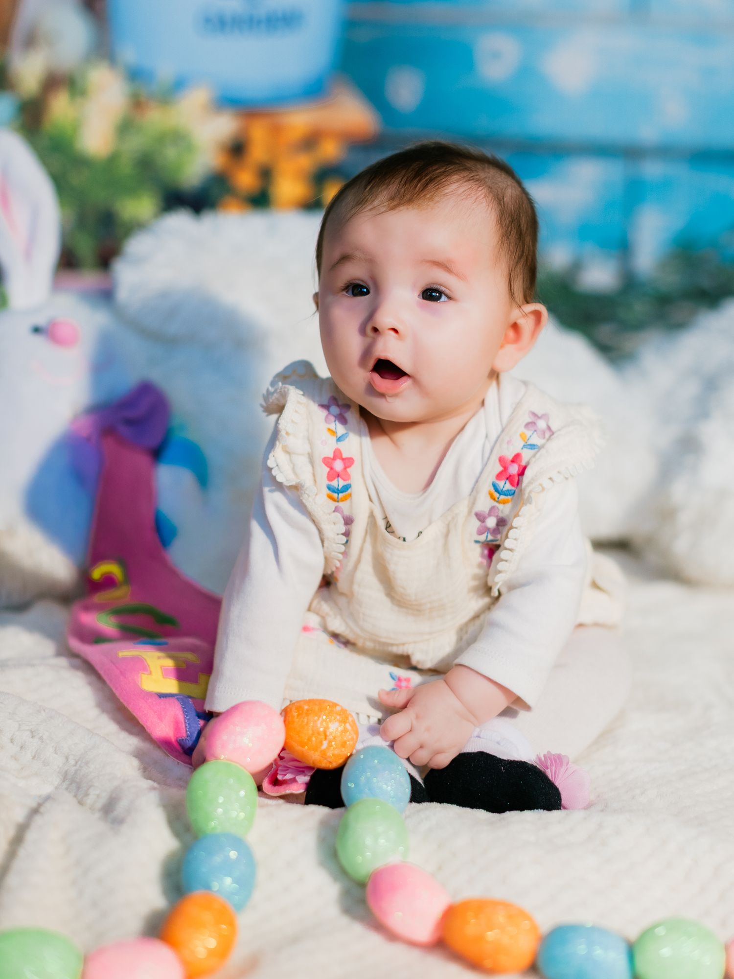 Adorable baby girl in white dress sitting on blanket surrounded by colorful Easter eggs during festive photo shoot.