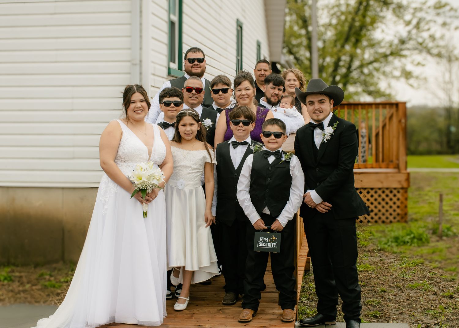 Wedding party in black and white attire with sunglasses poses outside a white chapel on a cloudy day.