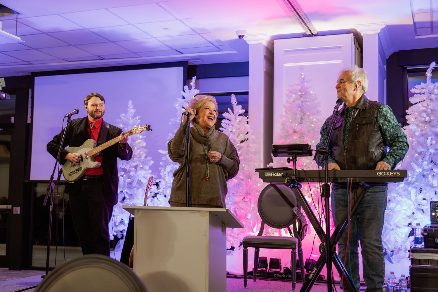 Musical performance setup with instruments and white Christmas trees against purple backdrop.