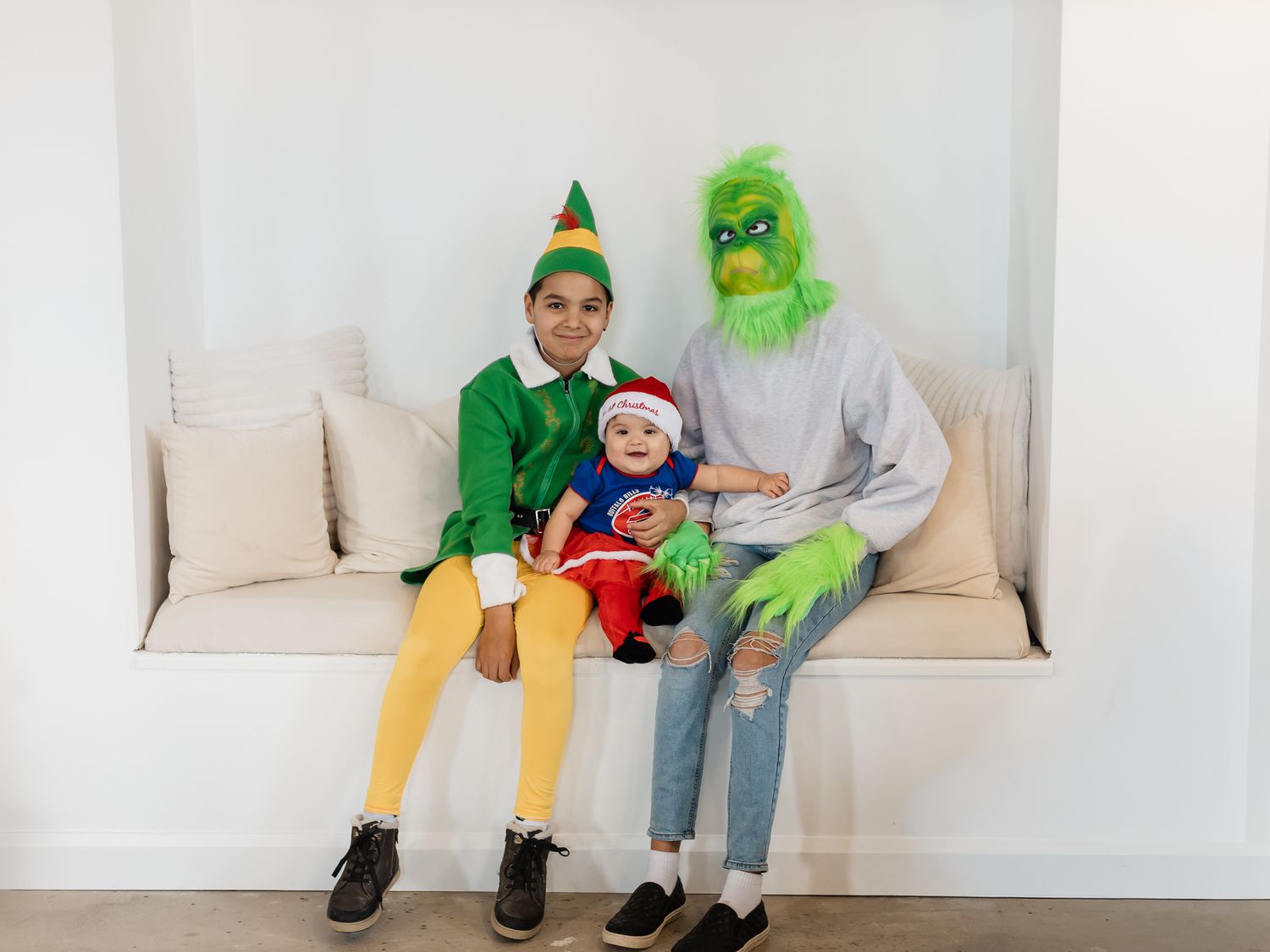 Three costumed characters including an elf and the Grinch pose together on a white bench.
