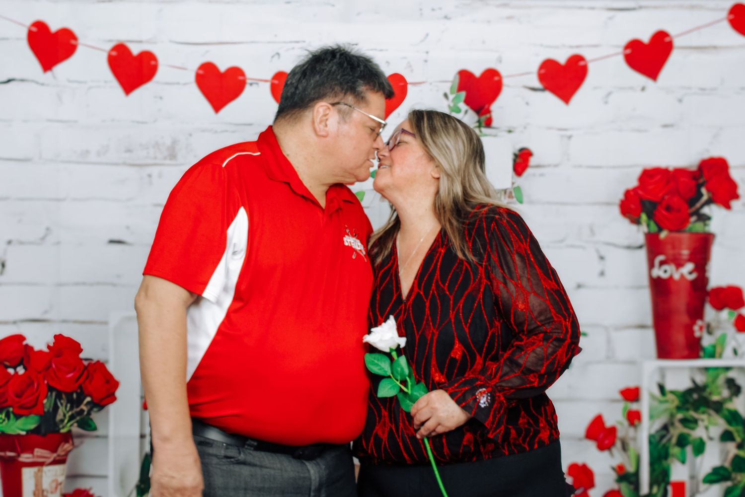 Romantic Valentine's day photo shoot with couple against white brick wall decorated with red hearts and roses.