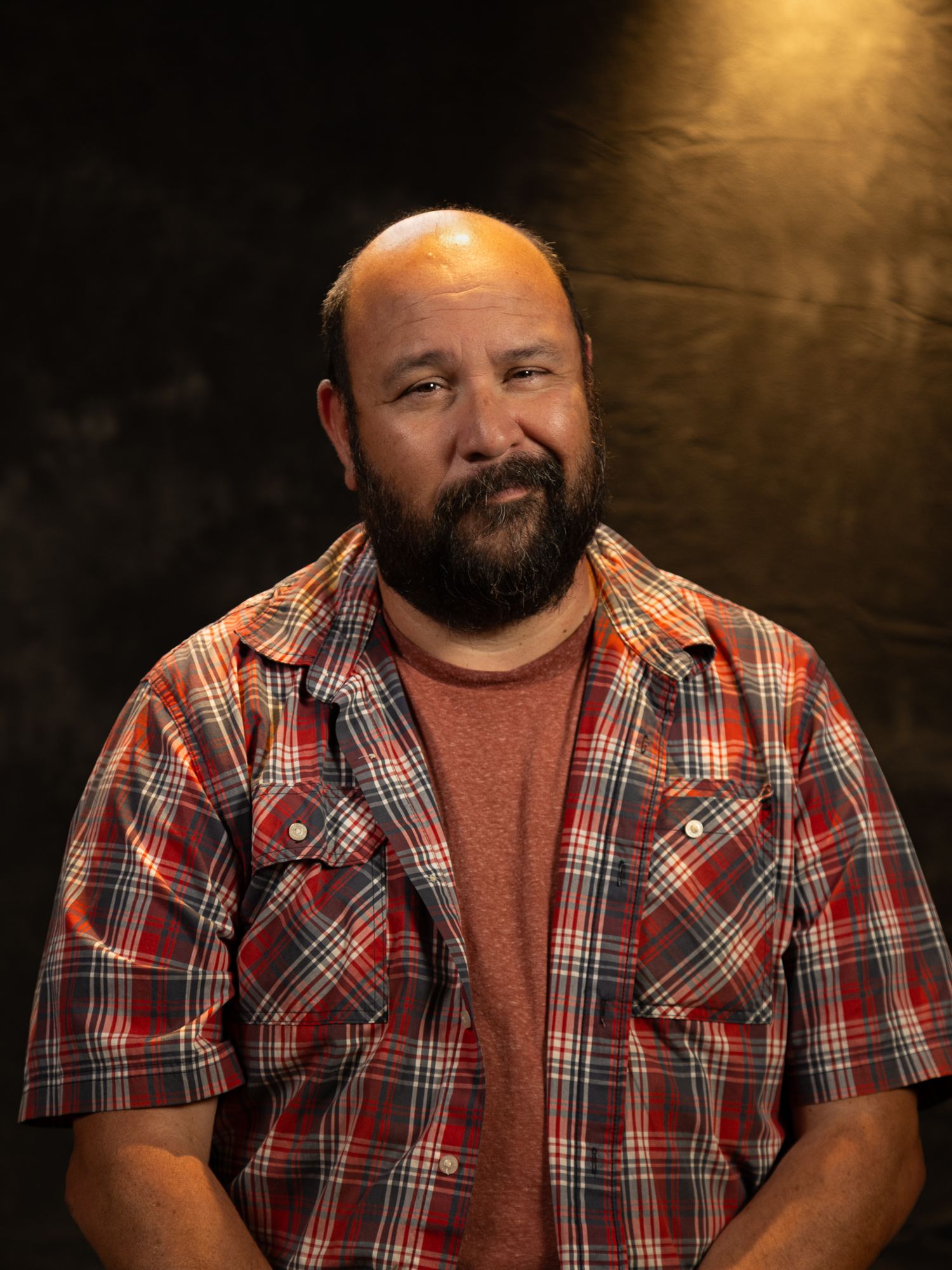 Studio headshot of person wearing plaid button-up shirt over rust colored tee.