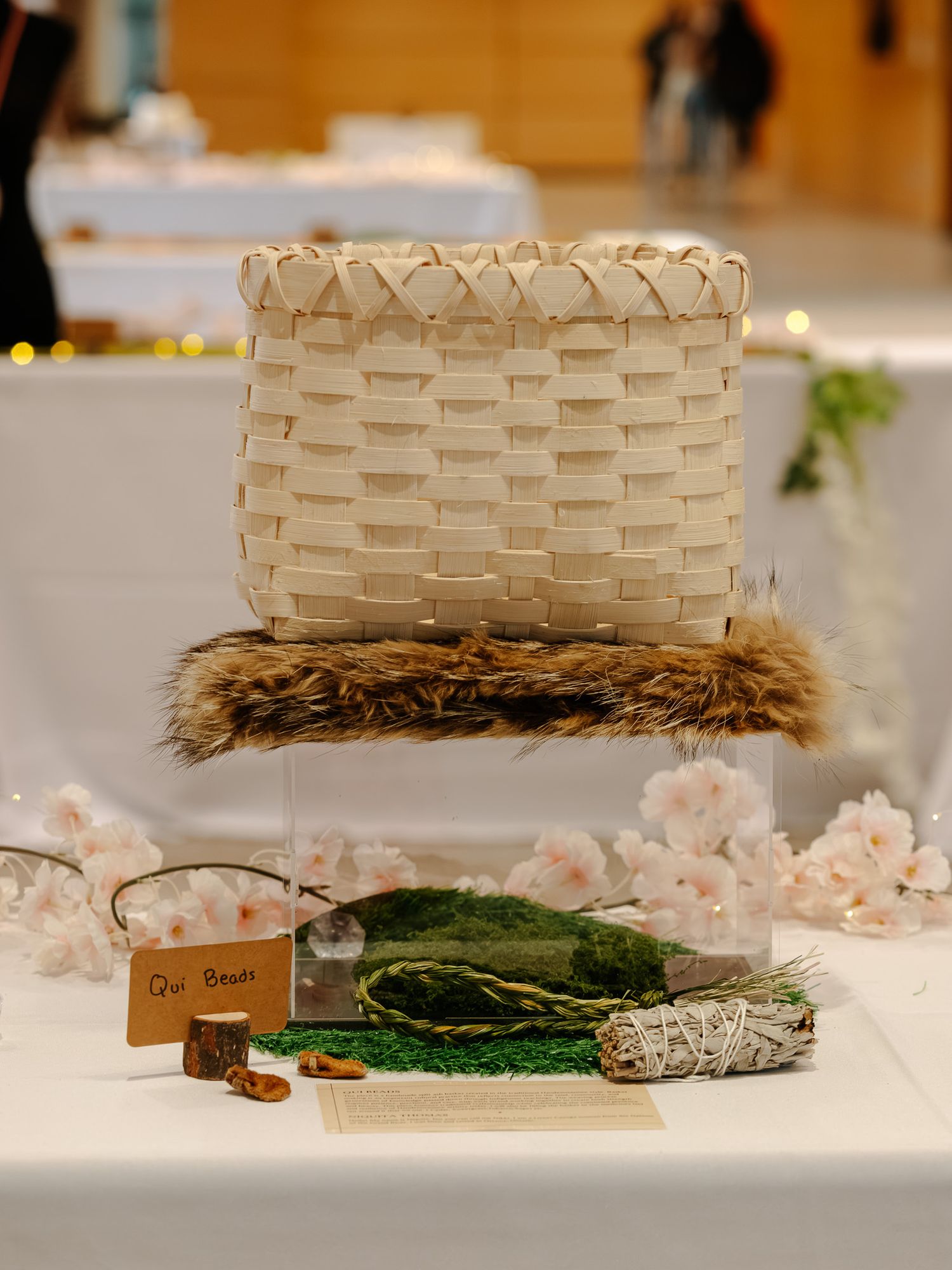 Woven basket display with fur trim, glass box containing moss and sage bundle on white table with cherry blossoms.