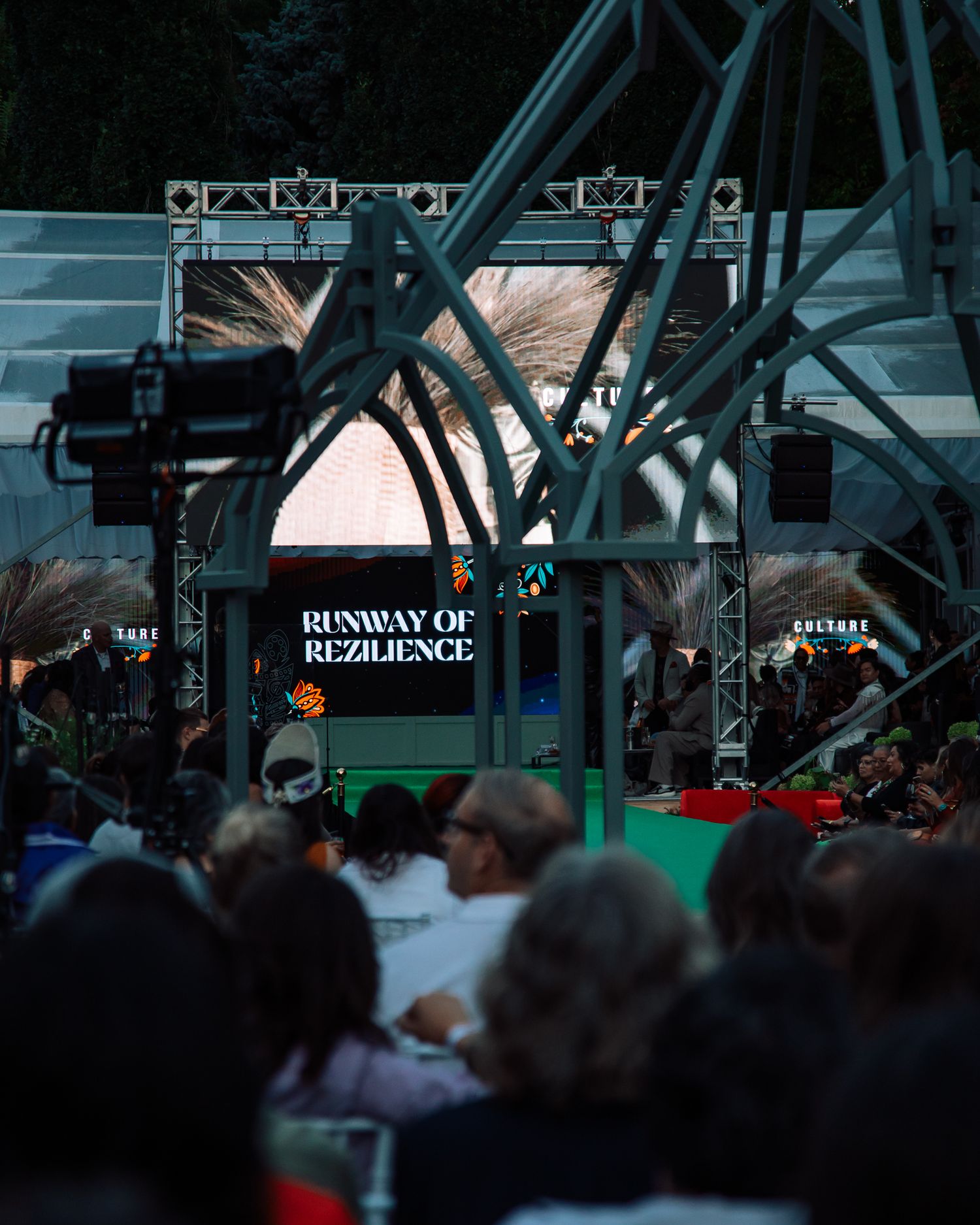 Crowd watches Runway of Rezilience Indigenous fashion show on illuminated outdoor stage at night.