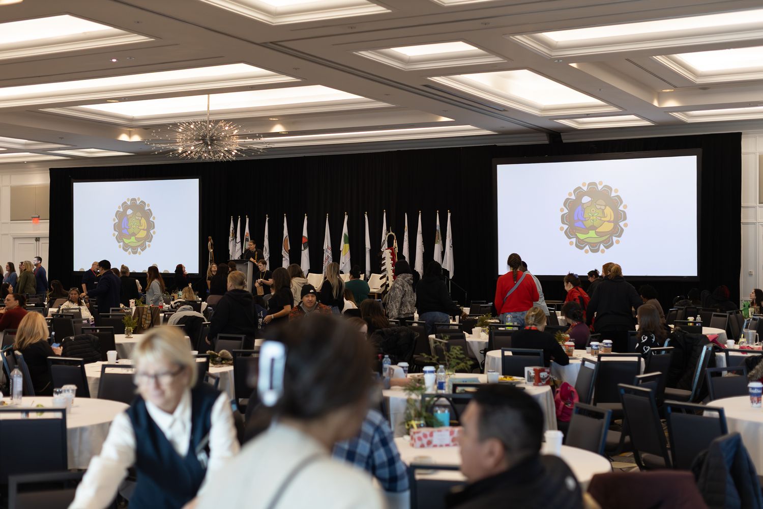 Large conference hall with attendees seated at round tables facing a stage with two large screens displaying an official seal.