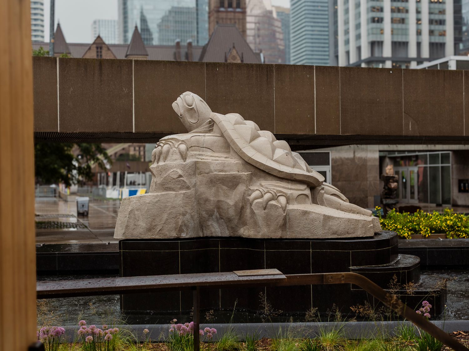 Large reclining marble statue in an urban plaza fountain with city skyscrapers visible in the background.