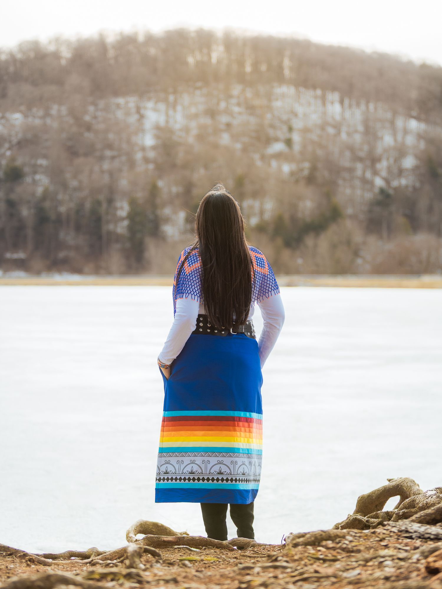 Woman in colorful Native American dress stands by a frozen lake with wooded hills in the background.